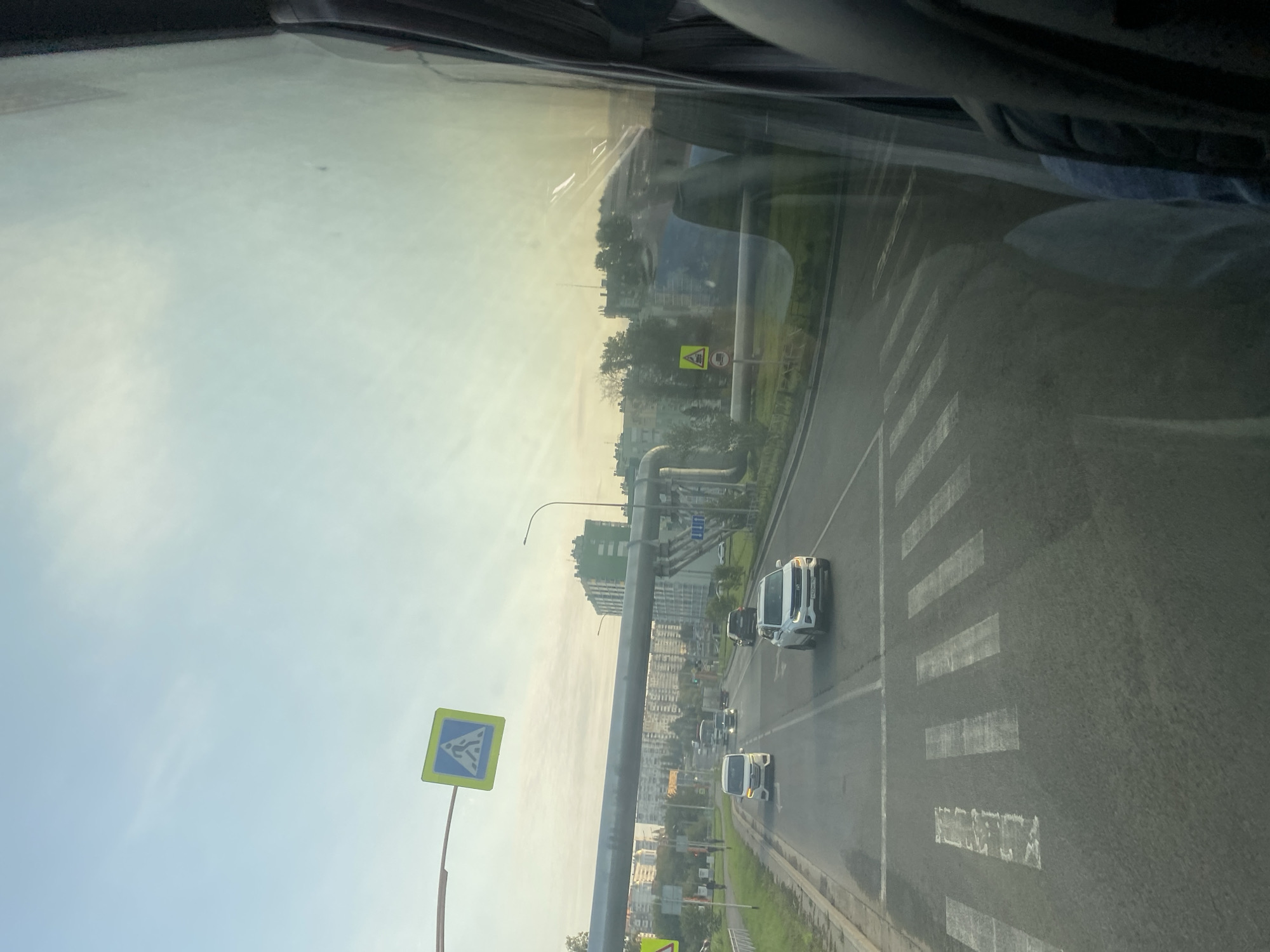 A slightly overcast day in Kemerovo, Russia, is depicted from the perspective of someone inside a vehicle, likely a bus, looking out through a window. The scene is dominated by a multi-lane road with a pedestrian crossing marked by white stripes. Several vehicles, including a white SUV, a white minivan, and a dark SUV, are visible on the road.
To the left of the road, there is a sidewalk, green grass, and then a row of apartment buildings and other structures. A large, elevated pipe, possibly for utilities, runs horizontally across the image, supported by poles. Traffic signs are present: a pedestrian crossing sign is visible on the left, and another warning sign is seen further down the road on the right. The sky above is pale blue with some haziness, and the sun appears to be low, casting a soft light. The window has some reflections, indicating the viewpoint is from indoors.