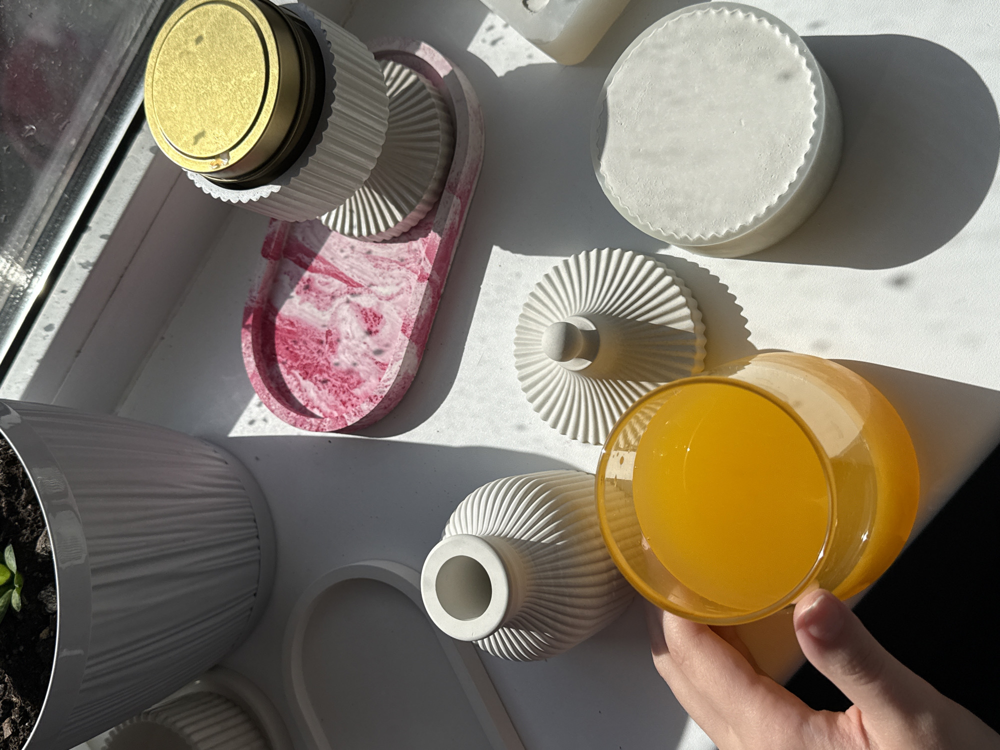 On a bright, possibly sunny day in an unknown city, several objects are arranged on a white windowsill. At the left, a white ribbed pot contains soil and a few small green plants. Adjacent to the pot are a couple of white ribbed bowls or bases, and above those is an oval tray with pink marbling. Next to the oval tray, a round container with a gold lid sits in a white ribbed base. To the right is a round white object with ridged sides. Further right and slightly below, a white ridged object lies next to an inverted cone-shaped object with a round knob on the end. Adjacent to it is a vase shape with a white exterior, next to which a hand holds a glass filled with orange liquid.