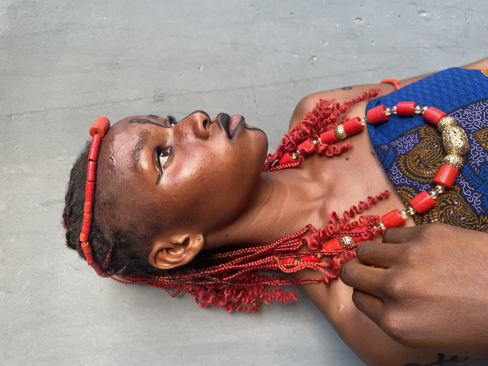 A high-angle, close-up portrait shows a person with dark skin lying on a grey, likely concrete, surface. The subject's head is angled upwards and to the left, with their gaze directed towards the upper left of the frame. Their face is adorned with striking black makeup designs, including a stylized sweep above the left eyebrow and outlining the eyes and lips.
A vibrant red beaded headpiece sits on their forehead, complementing the long, red braided and coiled hair extensions that cascade around their neck and shoulders. These extensions are intricately decorated with small red beads and gold-colored elements, forming a rich, ornamental collar. A prominent necklace of larger red barrel-shaped beads, interspersed with ornate gold-colored spherical beads, rests on their chest.
A piece of blue fabric with traditional yellow and black patterns is draped over their right shoulder and chest. A dark-skinned hand, possibly belonging to another person or the subject, is gently placed on the subject's upper chest near the necklace. The lighting is bright and even, suggesting daytime conditions without harsh shadows.
The image, set in Badagry, Nigeria, emphasizes cultural adornment and artistic expression, with the subject displaying a serene and contemplative expression. No text is visible in the image.