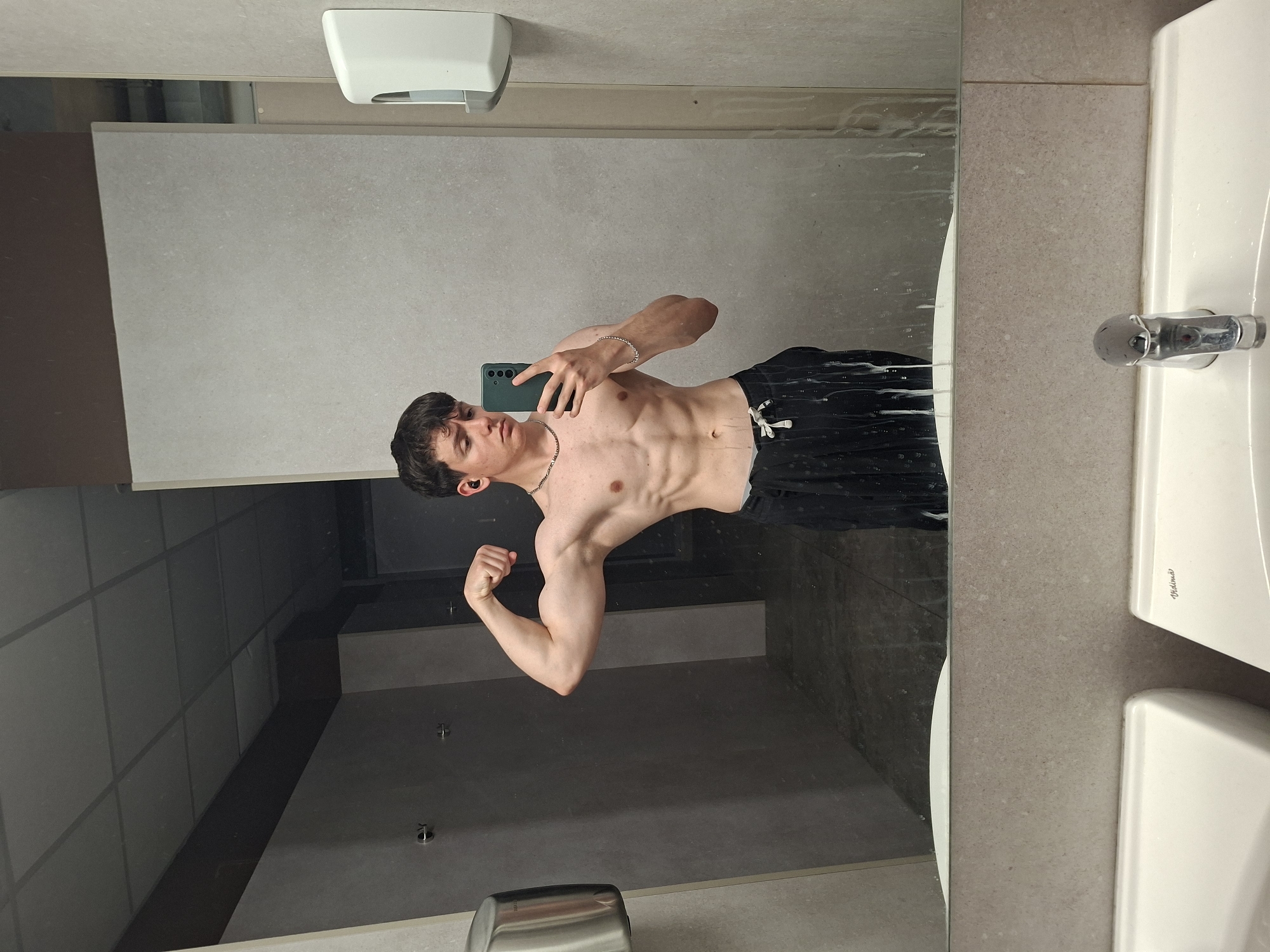 A young man with dark hair is captured in a mirror selfie, posing shirtless in what appears to be a public restroom or changing room. He is flexing his right bicep and showcasing his toned torso and abdominal muscles. He holds a dark green smartphone in his left hand, pointed towards the mirror to take the photo. He wears black shorts, a silver-colored chain necklace, and a bracelet on his left wrist, with a dark earbud in his left ear.
The setting features light brown or greyish tiled walls and a large mirror that reflects the room's darker interior, possibly showing ceiling panels. A white ceramic sink with a metal faucet is visible on the right side of the frame, along with a white hand soap dispenser mounted above the mirror and a metallic paper towel dispenser below. The mirror surface has white streaks and splashes, suggesting water or soap residue. The man's black shorts also appear to have similar droplets. The word "Vidima" is faintly embossed on the sink. The provided location context for this image is Plovdiv, Bulgaria.