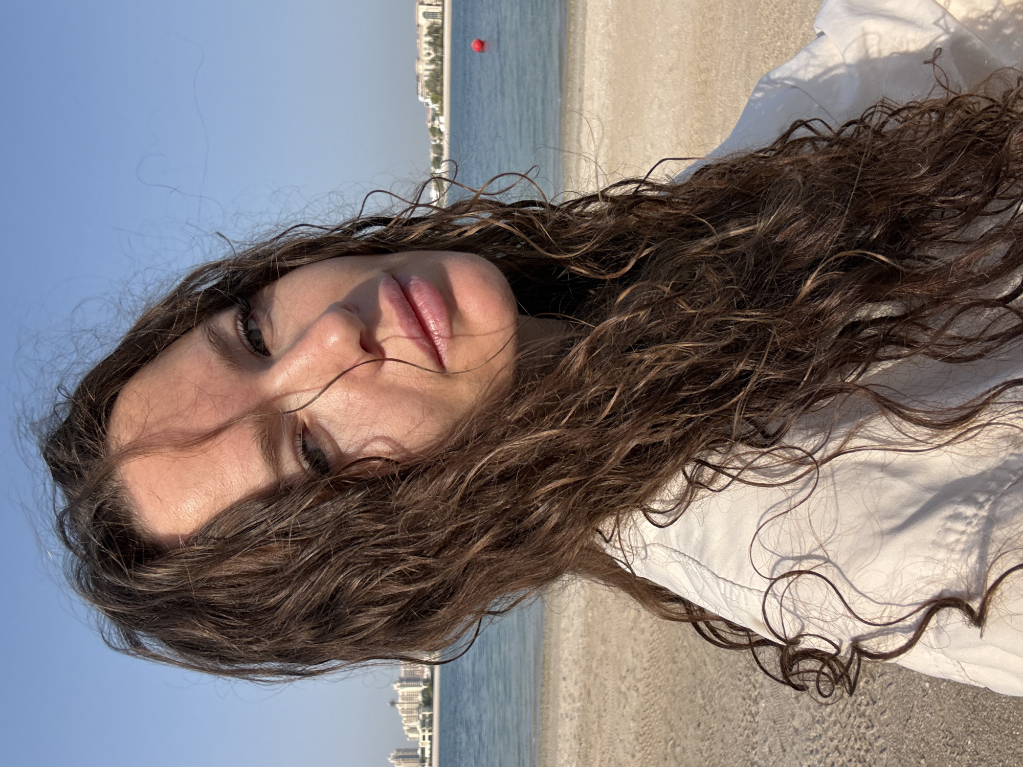 A close-up, selfie-style shot shows a woman with long, wavy brown hair on a beach in Dubai, United Arab Emirates. The woman is wearing a white shirt. Her face is partially obscured by her hair, but her eyes and lips are visible. The background shows a sandy beach, the blue water of the ocean, and buildings in the distance. A red buoy is visible in the water. The sky is clear and blue.