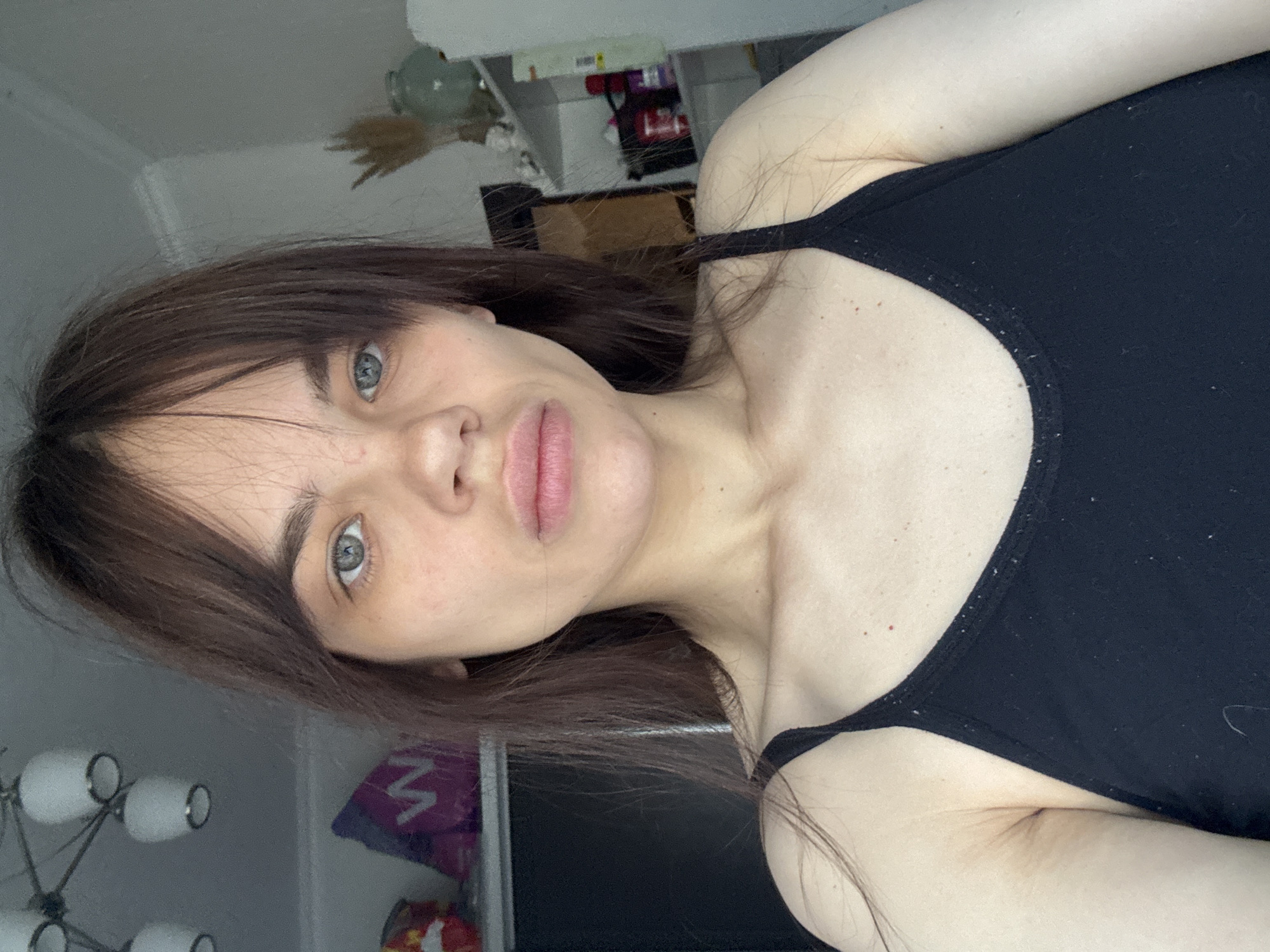 The image is a portrait-style photograph of a young woman, captured from a slightly upward angle and rotated 90 degrees clockwise.
The primary subject is a young woman with fair skin, dark brown, shoulder-length hair, and bangs. Her eyes are light-colored, appearing blue or grey-green, and she looks directly into the camera with a neutral, slightly contemplative expression. Her lips are naturally full and slightly parted. She is wearing a simple black tank top or camisole, which appears to have some light-colored lint or dust on it.
The setting appears to be an indoor room, likely a home, with various elements visible in the background, which is mostly out of focus. To the woman's left (when oriented correctly), a multi-bulb ceiling light fixture with white spherical shades is visible. To her right, shelves or a counter hold several obscured items, including a brown box and some dried, decorative foliage in a container. Below her left shoulder, a purple object or bag is partially visible, displaying a large partial letter that resembles a "W" or "M".
The lighting is soft and diffuse, suggesting indoor illumination or indirect daylight, and does not indicate a specific time of day. The woman is posing for the camera, possibly taking a selfie. There are no overt emotional expressions or specific activities taking place other than the direct gaze.
