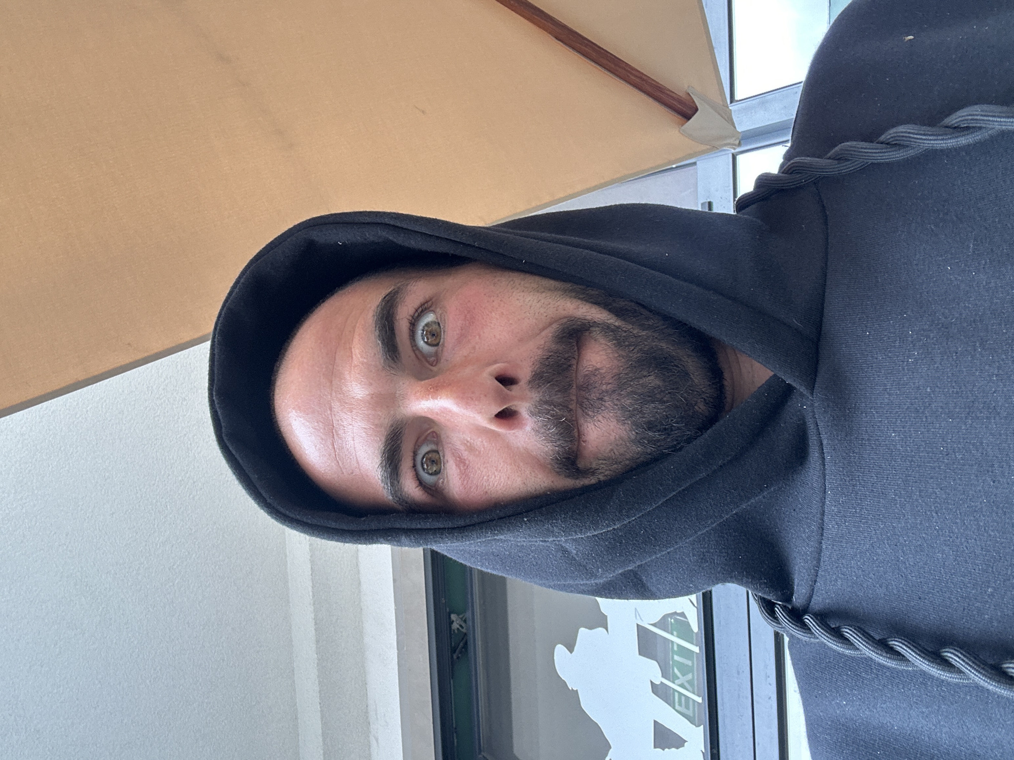 A selfie photograph taken by a man with light brown eyes, stubble, and a dark-colored hooded sweatshirt. He is positioned beneath a beige umbrella, partially visible in the upper left corner. Behind him, a glass door displays an "EXIT" sign and a human figure. The location is Budva, Montenegro. The photo appears to have been taken outdoors.