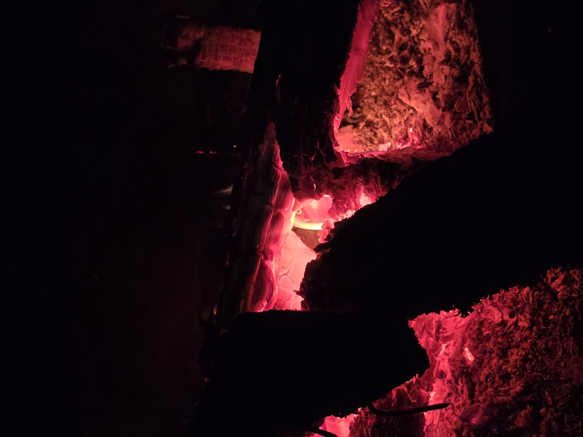 The image depicts a close-up view of glowing embers and charred wood in darkness. The scene is lit by the intense red and orange hues of the burning material, creating a stark contrast with the blackness surrounding it. There are no people visible, and the focus is solely on the remnants of a fire. The texture of the ash and burnt wood is prominent, with some pieces appearing to be partially intact logs or branches. The environment appears to be an outdoor setting, given the nature of the fire, and the location provided is Строитель-нефтяник Снт, Russia. There is no indication of any specific actions or interactions taking place in the image, beyond the ongoing process of burning and cooling.