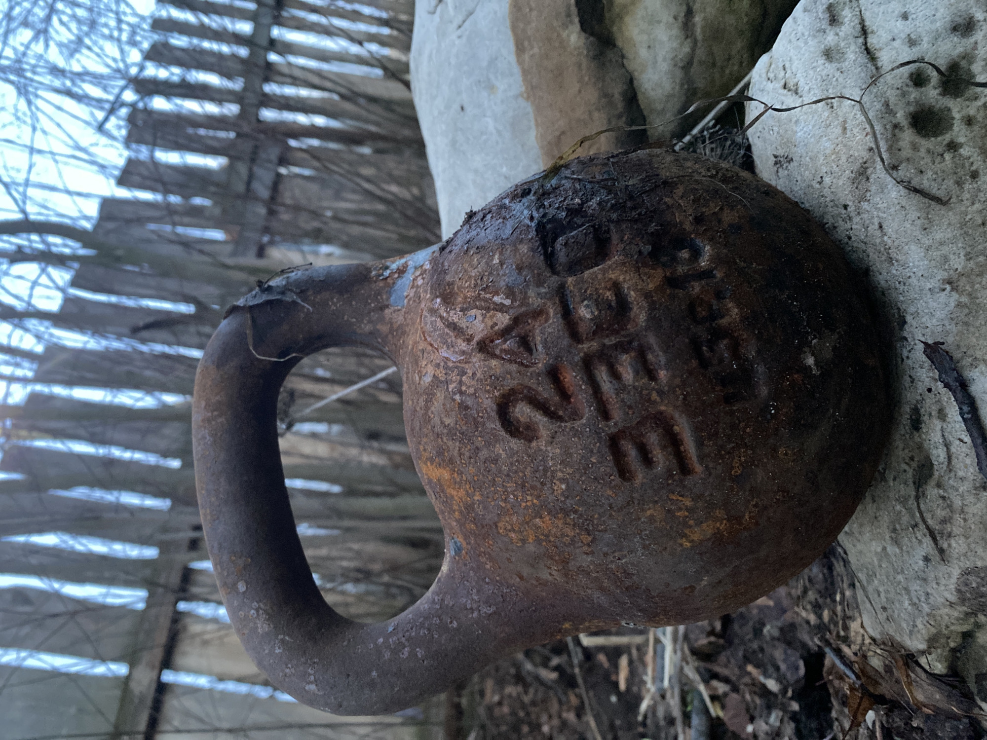 A rusty, 24-pound kettlebell rests on the ground near a light gray stone. A weathered wooden fence is visible in the background. The location is Ольховка, Russia.