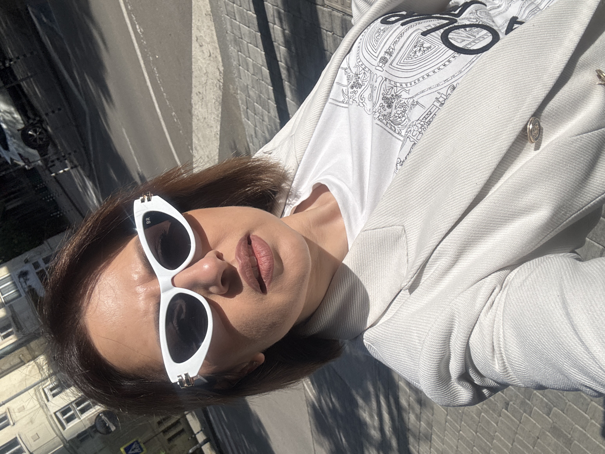A woman is posing in a selfie shot outdoors in Sofia, Bulgaria. She is wearing white sunglasses, a white T-shirt with a black design, and a light-colored blazer. Her hair is brown. In the background, there is a road with cars and a building with windows. The ground is paved with rectangular stones.