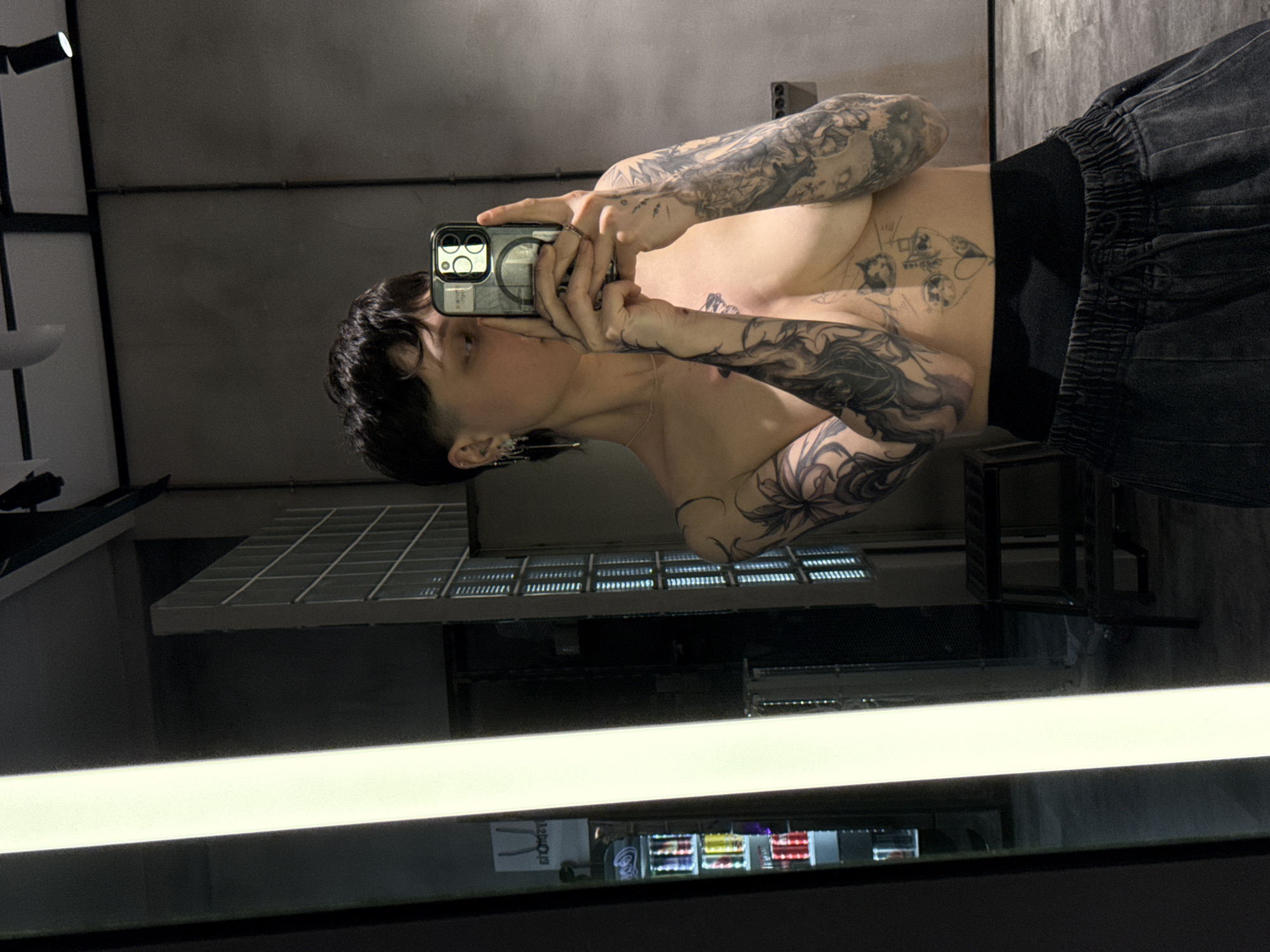 A person with short dark hair and extensive tattoos is taking a selfie in a mirror. The person is wearing dark-colored shorts with an elastic waistband and is shirtless. The background shows a room with a window featuring a metal grate and a cooler with beverages, reflected in the mirror. The cooler's reflection includes recognizable brands of beverages. The setting appears to be an indoor location, possibly a store or commercial space. The reflection in the mirror indicates the presence of a light fixture above. There is no immediate indication from the image that the location is in Moscow, Russia.
