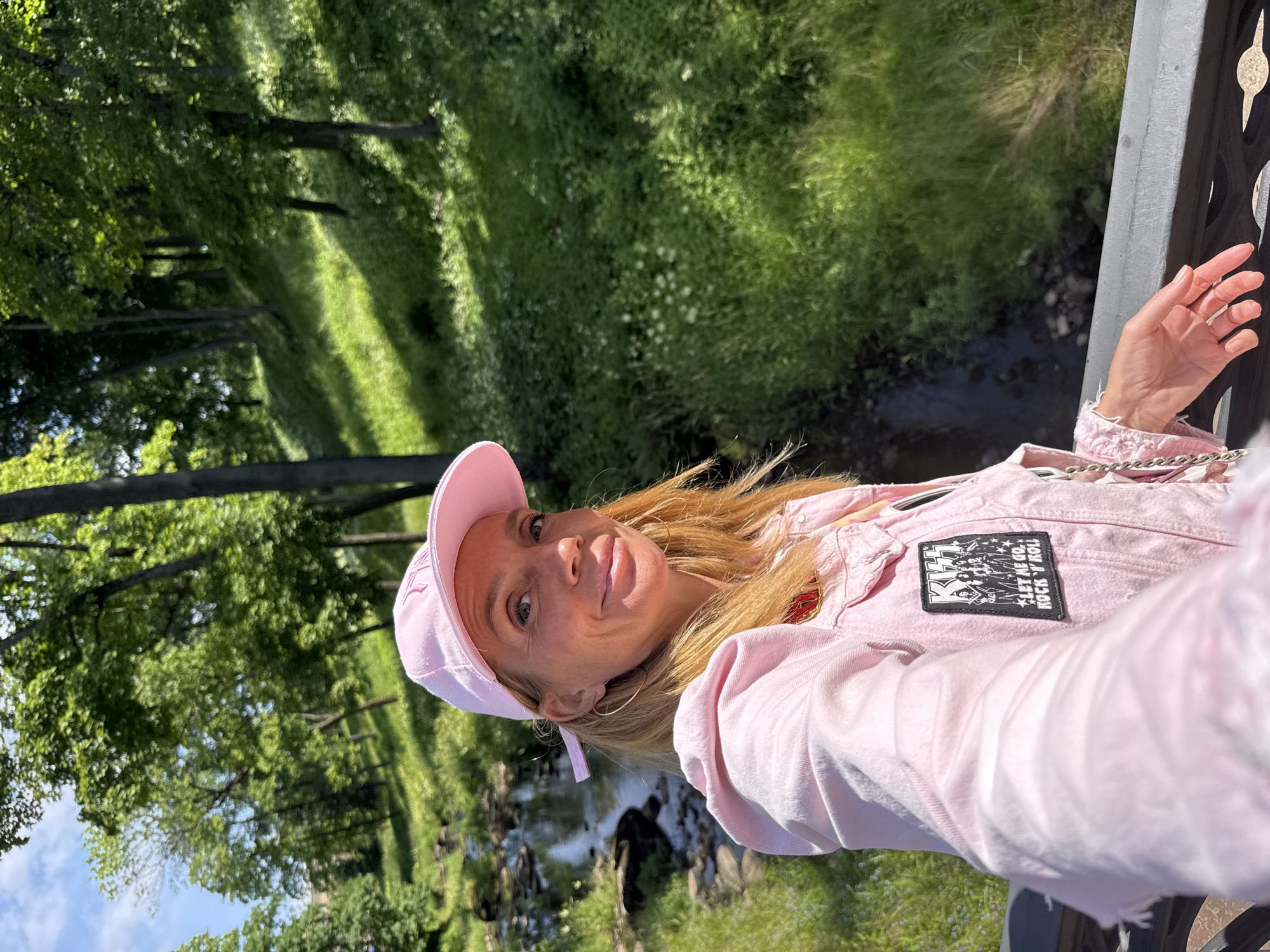 A blonde woman with fair skin and light freckles is taking a selfie outdoors. She is wearing a pink baseball cap, a pink denim jacket with a "KISS" patch that reads "LET ME GO, ROCK 'N' ROLL," and a gold hoop earring in her left ear. Her hair is light blonde and falls past her shoulders. She is smiling slightly as she looks at the camera. The background shows a wooded area with a stream running through it. The terrain is sloped and covered in green grass and trees. The location appears to be a park in the city of Lomonosov, Russia, possibly near the Grand Palace or its surrounding grounds, given the landscaping and natural elements. The lighting suggests it is daytime, with sunlight filtering through the trees.
