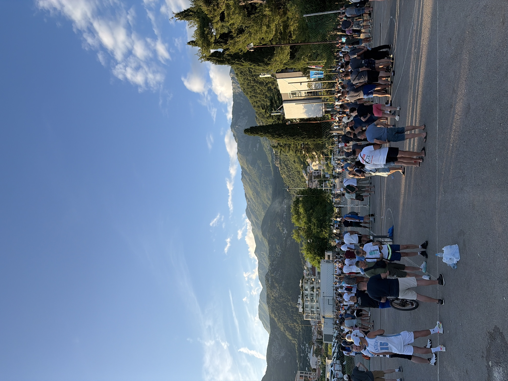 This media file captures an outdoor scene in Budva, Montenegro, likely during a public gathering in the late afternoon or early evening. The dominant features are a large crowd of people, imposing mountains, and a clear blue sky.
The scene shows numerous individuals standing on a paved surface, which appears to be a street, parking lot, or a sports court, judging by faint markings. They are lined up in multiple rows, facing an unseen event or direction to the right of the frame. Many individuals are casually dressed in t-shirts and shorts. Long shadows stretch across the ground, indicating the sun is relatively low in the sky, contributing to the warm, golden hour lighting.
In the background, a series of majestic, tree-covered mountains rise dramatically, suggesting a mountainous coastal or valley location. Nestled at the base of these mountains are several buildings with light-colored facades and red roofs, characteristic of Mediterranean architecture. Lush green trees, including tall cypresses, are scattered throughout the landscape and alongside the paved area, providing a natural backdrop.
The sky is a vibrant blue with scattered white, wispy clouds, suggesting fair weather. A chain-link fence runs along the edge of the paved area, separating the crowd from the mountainous terrain and some residential structures. On the ground near the crowd, a white plastic bag and a plastic water bottle are visible.
Visible text includes "ANTHONY" and the number "15" on the back of a white basketball jersey worn by a person in the foreground. Another white t-shirt has a red, partially obscured logo that resembles "Red Bull." The faint "SPAR" logo is visible on a white plastic bag on the ground. The overall atmosphere appears relaxed and expectant, with people gathered to observe an event.