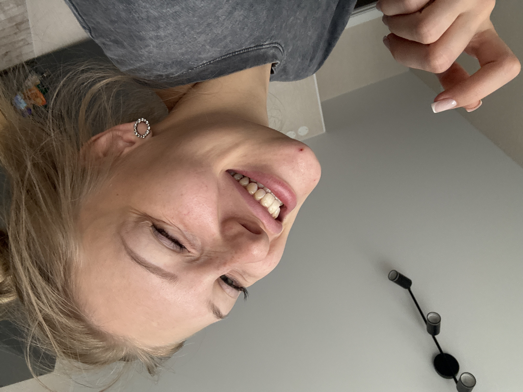 A young woman with blonde hair is pictured from the chest up, smiling and looking towards the camera. She is wearing a gray t-shirt and a circular earring with small stones on her right ear. Her left hand is raised, with her fingers in a pinching gesture. She has a mole on her chin. In the background, there is a ceiling with a black light fixture and a wall. The image is taken indoors. There is no discernible connection to the city of Perm, Russia.