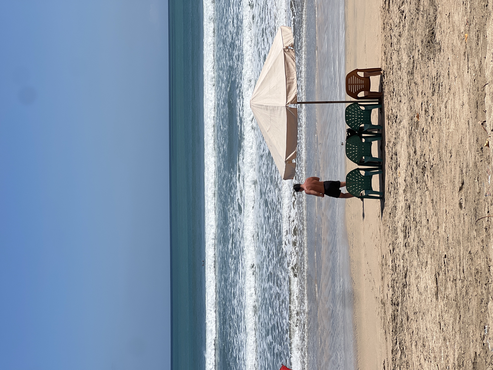 The image depicts a serene beach scene in Kuta, Indonesia. A man stands facing the ocean, his back to the camera, wearing black shorts. In the foreground, a beige umbrella provides shade over four plastic chairs: three green and one brown. The ocean stretches out to the horizon, with gentle waves breaking on the sandy shore. The sky is clear and blue, indicating a sunny day.