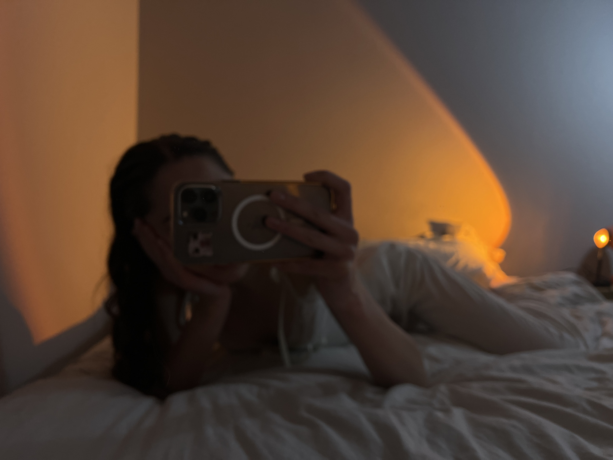 A woman with dark hair is lying on a bed in a room, obscured by a cell phone she is holding up to take a picture. She's wearing light-colored pajamas. The room has a warm, diffused light, with a noticeable streak of light across the wall behind her. There is a small lamp to the right of her bed. There is no information in the image to suggest this is in Ternopil, Ukraine.