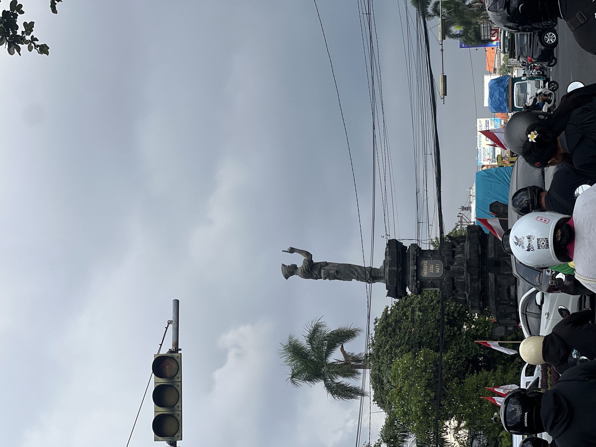 In Denpasar, Indonesia, a traffic scene unfolds under a cloudy sky. In the foreground, several people are seated on motorcycles, their faces obscured by helmets. One helmet, worn by a person on the far left, is black with the "HONDA" logo visible. To their right, another person wears a beige helmet. The central figure in the image is a statue of a soldier, standing on a stone pedestal, pointing one finger upwards. The pedestal has an inscription that reads "MERDEKA ATAU MATI," meaning "Independence or Death" in Indonesian. Behind the statue, lush green trees and a palm tree are visible. A traffic light hangs overhead, with the red light illuminated. In the background, various vehicles, including cars and trucks, are present, suggesting traffic congestion. Several Indonesian flags are also displayed, some attached to poles and others on vehicles. The overall scene depicts a busy intersection with a prominent historical monument.