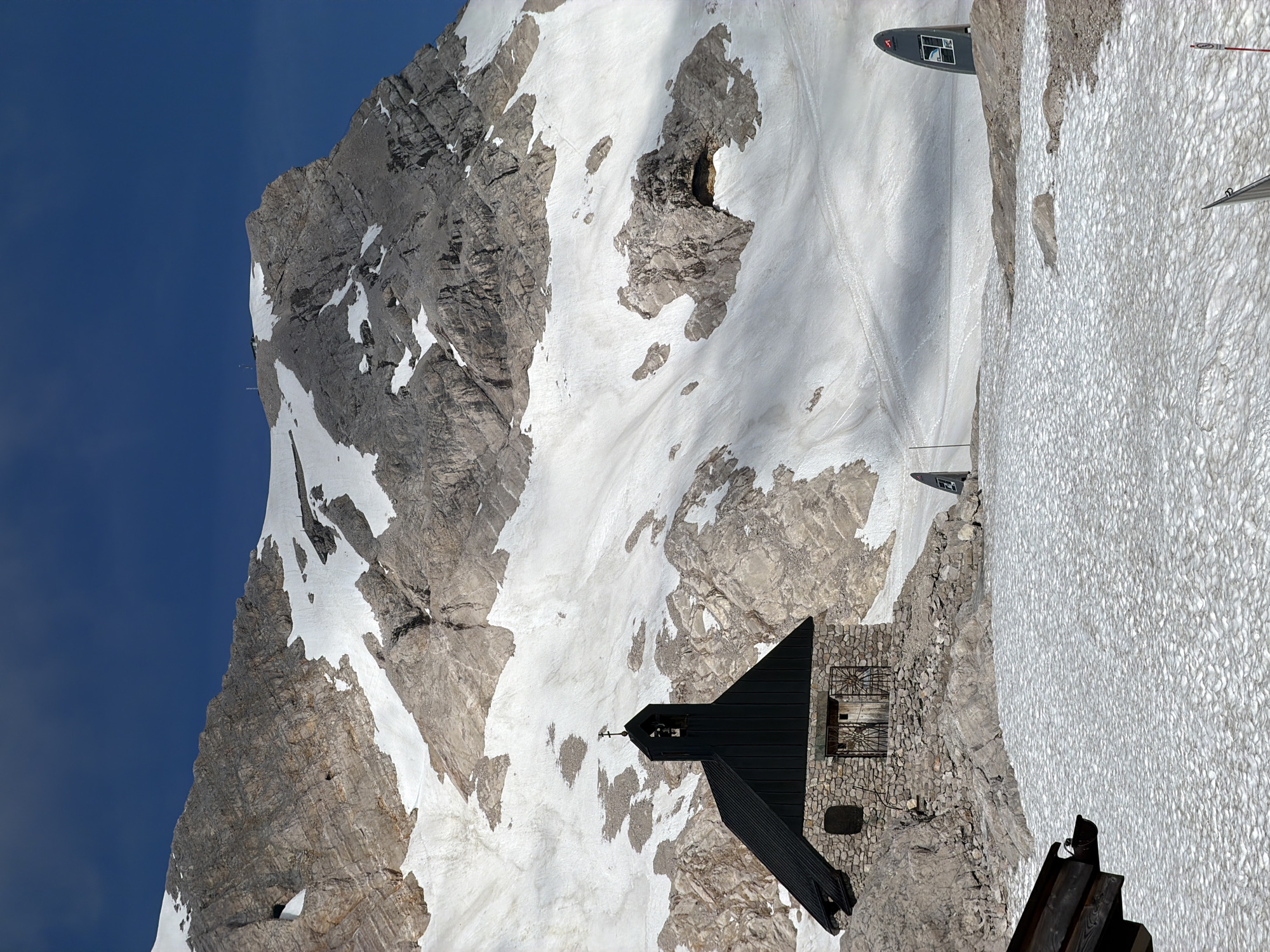 The image depicts a snowy mountain landscape with a small stone building topped with a dark roof, featuring a steeple. The building appears to be a chapel or a small mountain hut. Behind it, a vast, rugged mountain face is visible, partially covered in snow. The sky above is a deep blue. In the foreground and middle ground, there are patches of snow and rocky terrain. Two dark, curved signs are present on the right side of the image. The scene suggests a high-altitude, remote location, likely in the Bavarian Alps near Garmisch-Partenkirchen, Germany, given the context. There are no people visible in the image. The environment is characterized by rock, snow, and a clear sky.