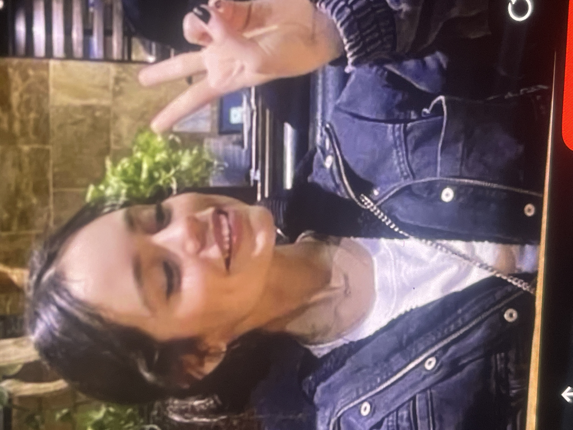 A vertical, medium shot captures a young woman smiling and making a peace sign with her right hand, suggesting a cheerful and playful moment. The scene is likely indoors or in a covered outdoor area in Poltava, Ukraine.
The woman, with dark hair parted on the side, is looking directly at the camera with a wide, open-mouthed smile, her face expressing happiness. She is wearing a dark blue denim jacket over a white top, part of which is visible around her neck and chest. A thin silver chain, possibly a necklace or part of a bag strap, is visible across her chest. Her right hand is raised, with the index and middle fingers forming a "V" for peace, and her fingernails are painted dark. A black bag strap with metallic links is visible draped over her shoulder.
The background features a tiled wall in shades of brown and gold, with varying textures. Below the tiled wall, green foliage, possibly potted plants, adds a touch of nature to the setting. Above the foliage, vertical metallic elements, resembling pipes or decorative bars, are visible. The lighting suggests an indoor or evening setting with artificial illumination.
Visible screen artifacts include a white circular loading icon in the top right corner and a white arrow icon at the bottom right, against a red and black interface strip on the right edge, indicating the image is a screenshot or photo of a screen. There is no discernible text within the depicted scene itself.