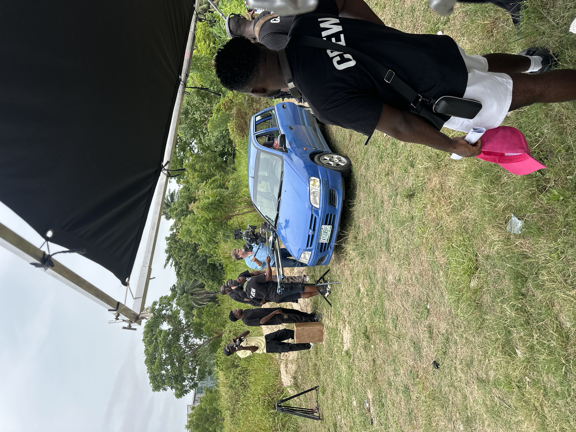 A film crew is working on location. A blue car is the central subject, positioned on a grassy patch with trees in the background. A cameraman stands in front of the car with a camera mounted on a tripod. Other crew members are present, some wearing "CREW" t-shirts. A black shade is visible on the left side of the frame, supported by a metal structure. One crew member is holding a pink hat and white paper. The scene is set outdoors, likely in Lagos, Nigeria, based on the car's license plate.