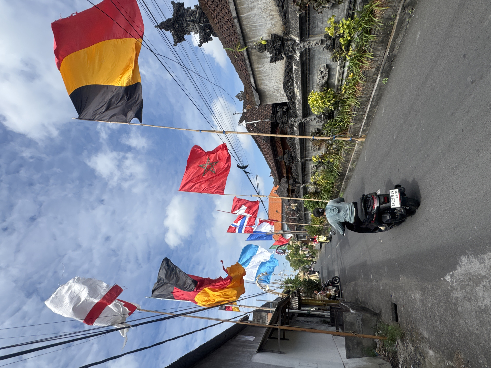 In Kuta, Indonesia, a street scene unfolds under a cloudy sky, adorned with a series of national flags flying from bamboo poles lining the roadway. The flags, including those of Germany, Belgium, Norway, and Morocco, are prominently displayed against a backdrop of traditional Balinese architecture. A person on a black scooter navigates the asphalt road, followed by another individual on a yellow scooter further ahead. The sides of the street feature buildings with tiled roofs and intricate stone carvings, interspersed with lush green foliage. The atmosphere suggests a festive or celebratory occasion, possibly a cultural event or the welcoming of foreign visitors.