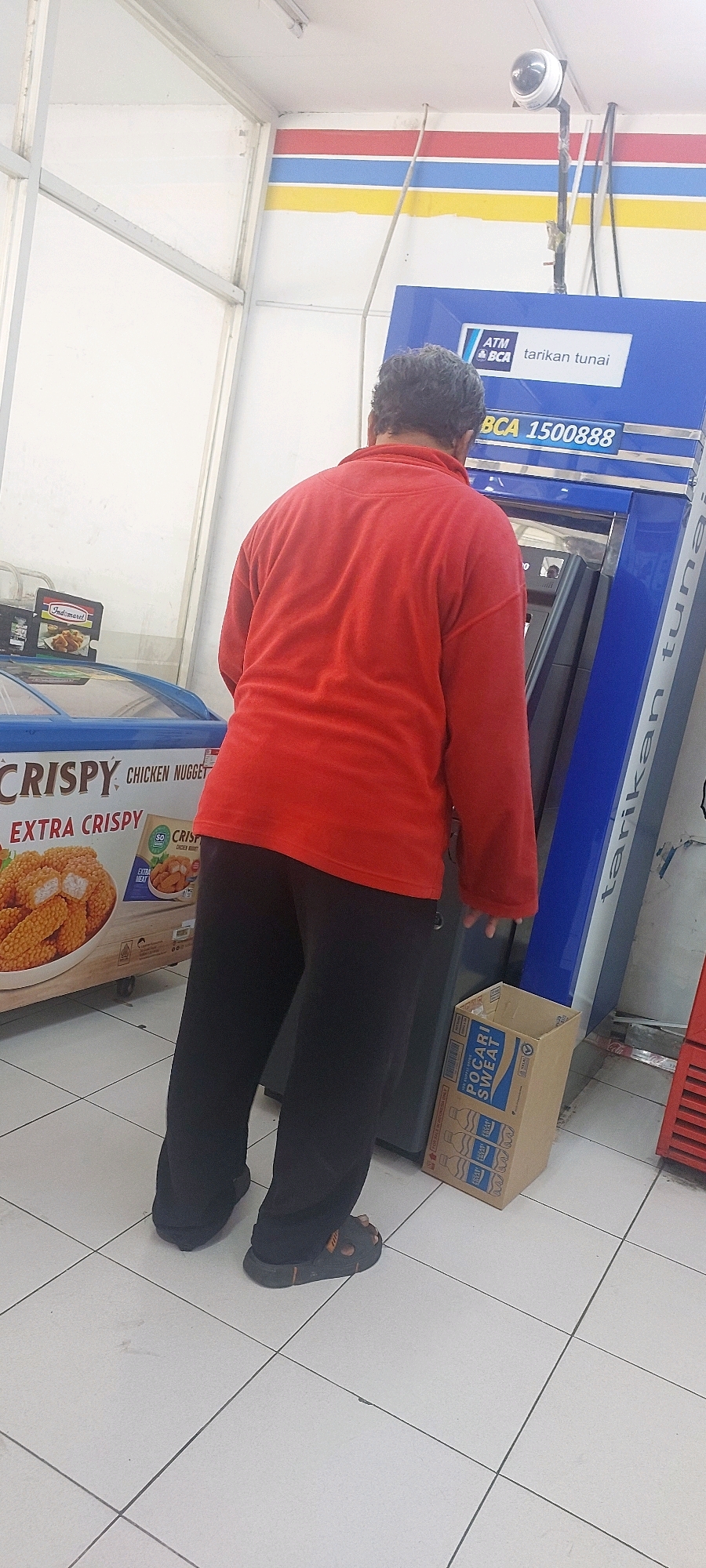 A person wearing a red long-sleeved shirt and black pants is standing in front of a blue ATM machine with "BCA 1500888" displayed. The ATM is labeled "ATM & BCA tarikan tunai" (meaning ATM & BCA cash withdrawal). To the left of the ATM, a person is standing next to a freezer case with a sign that says "CRISPY CHICKEN NUGGET EXTRA CRISPY". A cardboard box labeled "POCARI SWEAT" is on the floor to the right of the ATM. The scene appears to be inside a convenience store or a similar establishment, with white tiled flooring and a striped wall in the background. The lighting suggests an indoor environment, possibly with fluorescent lights overhead. The camera is positioned at eye level, capturing the scene from behind the person at the ATM. The environment looks like a typical Indonesian convenience store, possibly in the city of Semarang, based on the ATM branding and common store layout.