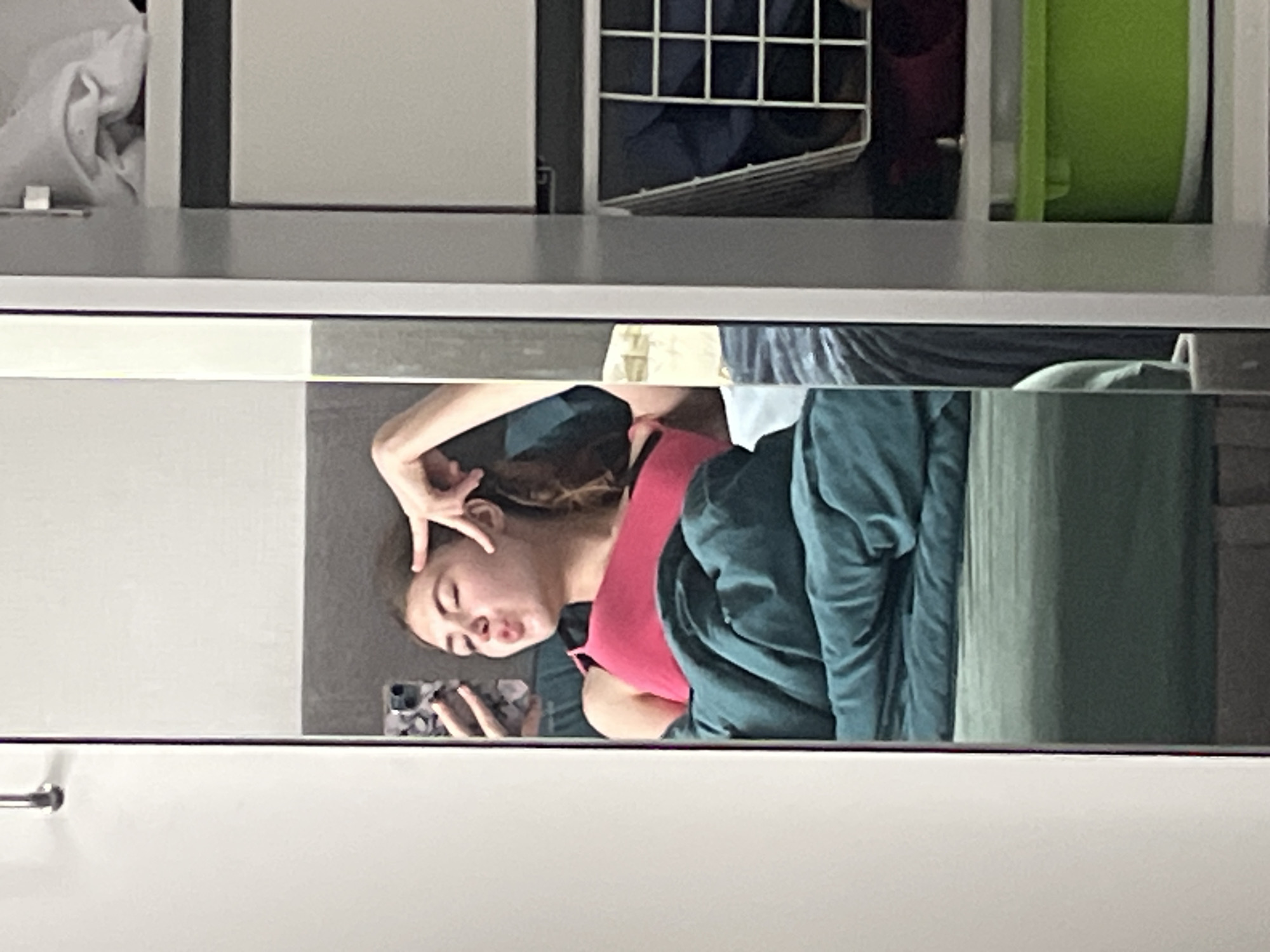 A young woman with brown hair is taking a selfie in a full-length mirror. She is lying in bed, covered with a dark teal comforter. She is wearing a bright pink tank top and is puckering her lips while holding her phone in her left hand and using her right hand to gesture near her temple. The mirror is positioned in front of a wall with some clothing storage units to the right. The scene appears to be indoors, likely in a bedroom. The lighting is soft, suggesting it could be natural light from a window or a well-lit room. The background of the mirror shows a hint of a window or wall behind the bed. There is no identifiable information in the image that would suggest it is located in Moscow, Russia.