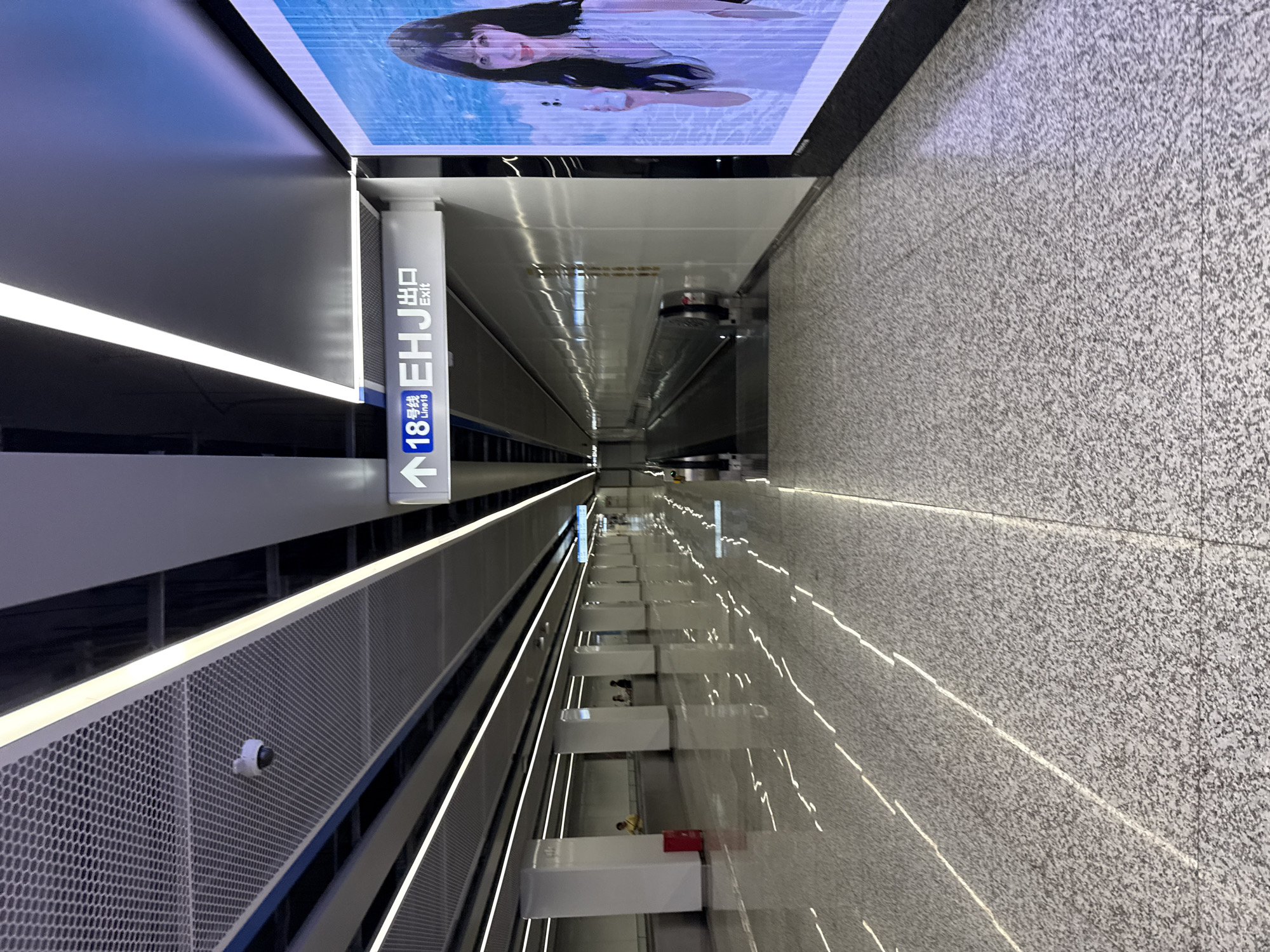The image shows an interior view of a modern subway station, likely in Haizhu District, China, given the presence of Chinese signage. The scene is characterized by a long, well-lit corridor with a moving walkway on the right side. The floor is a polished, speckled gray material, reflecting the linear light fixtures on the ceiling and the patterned lights on the floor.
In the foreground, the corridor extends into the distance. To the left, there are several white support columns, some with red trash receptacles. A few individuals are visible in the background, appearing to be waiting or walking. One person in a yellow shirt is near the red bin.
Above the moving walkway, a directional sign is prominently displayed. It features an upward arrow, the number "18" with Chinese characters indicating "Line 18," and the English word "Exit" preceded by "EHJ." To the far right of the image, a large digital billboard displays an advertisement featuring a young woman holding a smartphone.
The ceiling is a combination of a white perforated panel and a darker material, with recessed linear lighting that contributes to the bright and clean atmosphere of the station. The overall impression is one of efficiency and contemporary design.