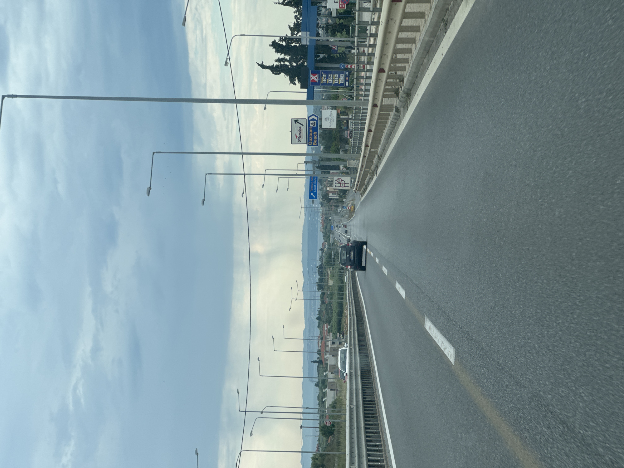 The image depicts a highway scene in Nea Moudania, Greece. The photograph is taken from a vehicle moving along the right lane of the highway. The road is grey asphalt marked with white lines. A dark-colored car is visible ahead in the same lane. To the left of the highway, there are multiple streetlights lining the road. Further beyond, there is a landscape with greenery, buildings, and hills in the distance. The sky is partly cloudy with a light blue hue. Gas stations and road signs are visible at a distance.