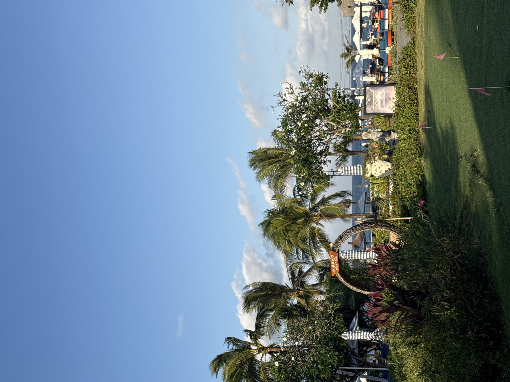 The image depicts a sunny day in Kuta Selatan, Indonesia, with a clear blue sky and scattered white clouds. In the foreground, a neatly manicured putting green is visible with three pink flags marking holes. Surrounding the green is lush vegetation, including tall palm trees swaying in the breeze, a prominent woven archway with a sign reading "Ayodya Resort Bali," and various bushes and flowers.
In the background, several beach umbrellas and lounge chairs are set up on the sand, suggesting a resort or beach area. The ocean is visible in the distance, with a calm surface. A few people are scattered among the lounge chairs, appearing to be relaxing. The overall scene conveys a serene and tropical atmosphere typical of a beachfront resort.