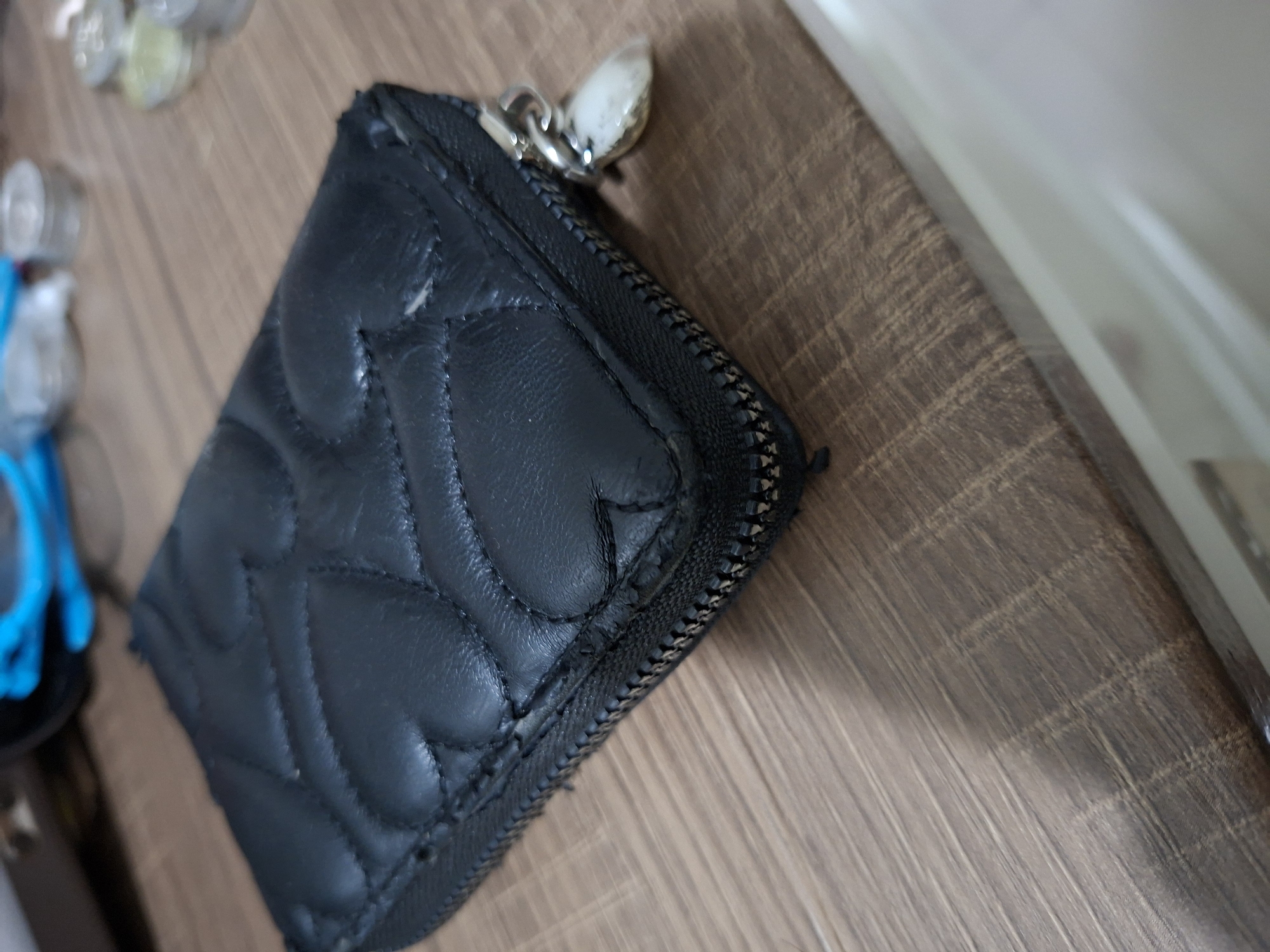 A black quilted wallet with a heart pattern is placed on a wooden surface. The wallet has a zipper closure and a silver charm hanging from the zipper pull. The background shows a blurry image of some containers and a blue object. The wooden surface appears to be a tabletop or desk. This image is taken from a high angle, looking down at the wallet. The lighting suggests an indoor setting. The overall scene is uncluttered and focuses on the wallet.