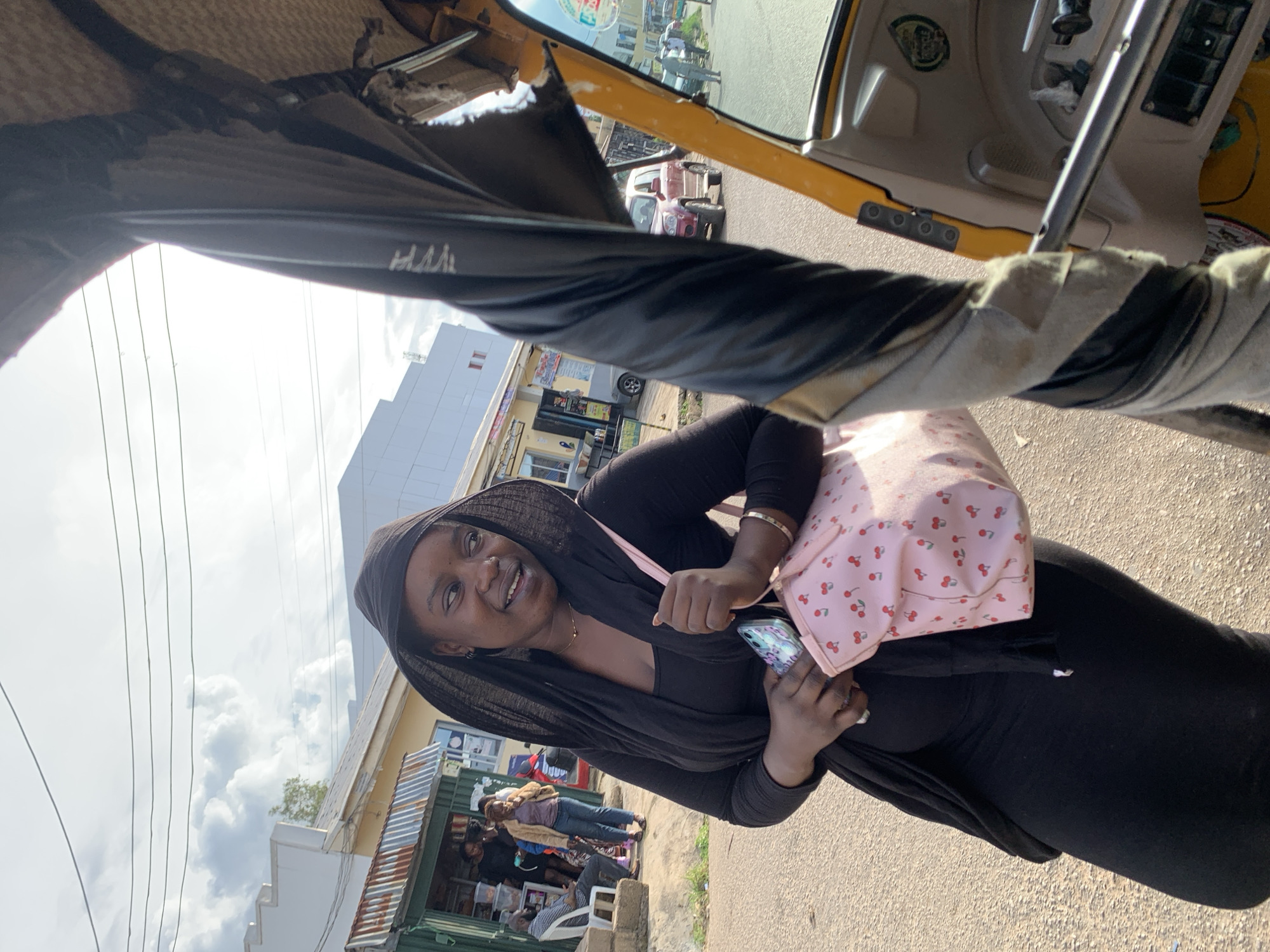 A woman wearing a black hijab and black dress stands in front of a row of shops. She is holding a light pink purse with a cherry pattern and a smartphone. In the foreground, the interior of a yellow auto-rickshaw is visible, with its canopy partially obscuring the woman. The background shows a street with other vehicles and people. The scene appears to be in Jos, Nigeria, given the architecture and context.
