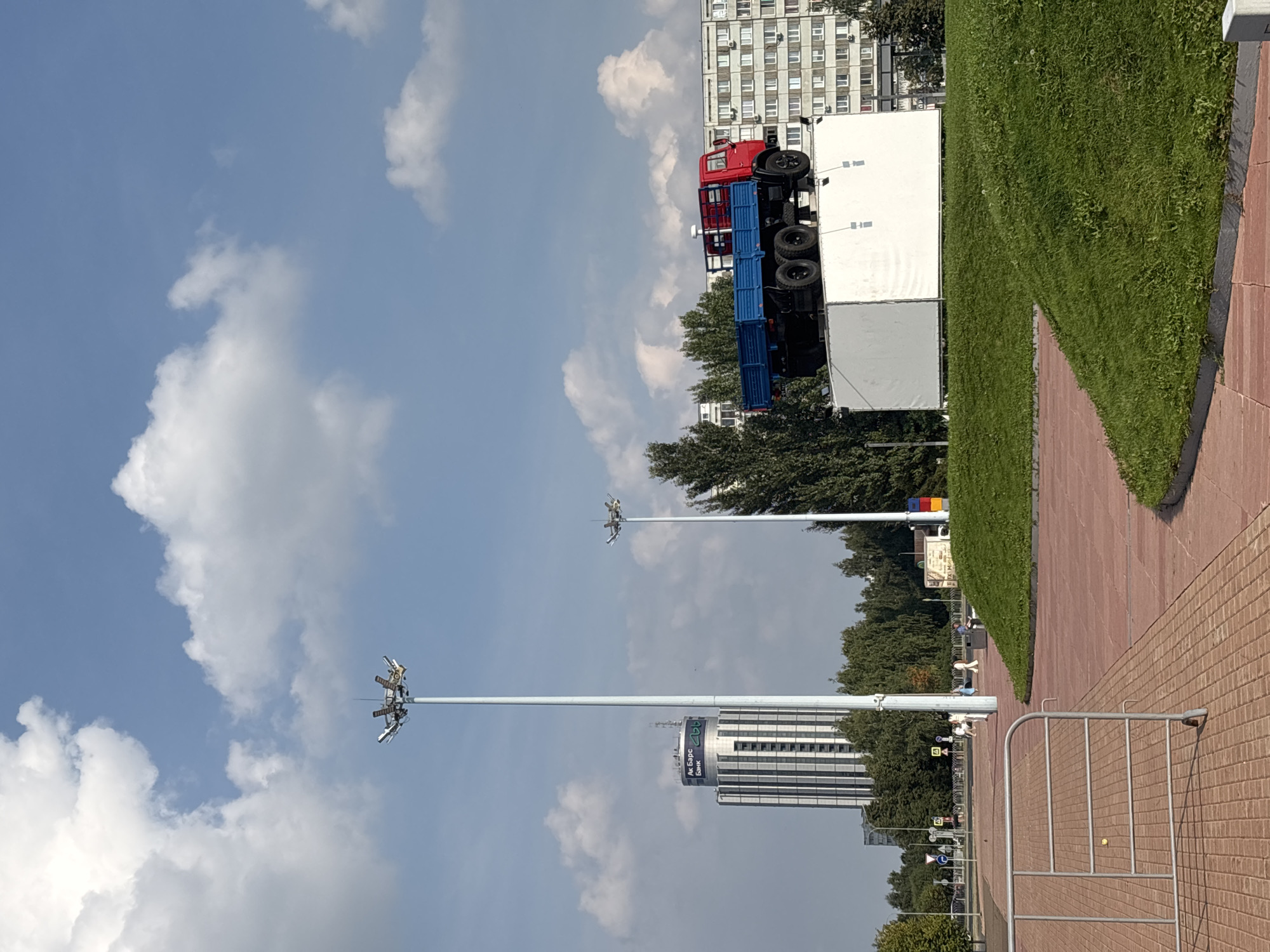 An outdoor daytime shot captures a monument featuring a red and blue flatbed truck positioned atop a large white structure. The truck is a prominent subject, with its red cab and blue flatbed clearly visible. The white structure appears to be a platform or base, possibly for display or artistic purposes, and is situated on a grassy mound.
In the background, several elements indicate an urban environment. A tall, modern building with a distinctive facade, likely an office building or bank, is visible to the left. Adjacent to it, a tall lamppost with multiple lights stands prominently. Further to the right, a row of apartment buildings with numerous windows lines the horizon. Trees and foliage are interspersed throughout the scene, suggesting a park or public space. The sky is blue with scattered white clouds.
A paved walkway or plaza occupies the foreground, with a metal barrier to the left. A few people are seen in the distance, walking on the paved area, but they are not the focus of the image. The overall scene appears to be a public square or area in the city of Naberezhnye Chelny, Russia, given the architectural style and the likely presence of the "Ak Bars Bank" signage on the prominent building.