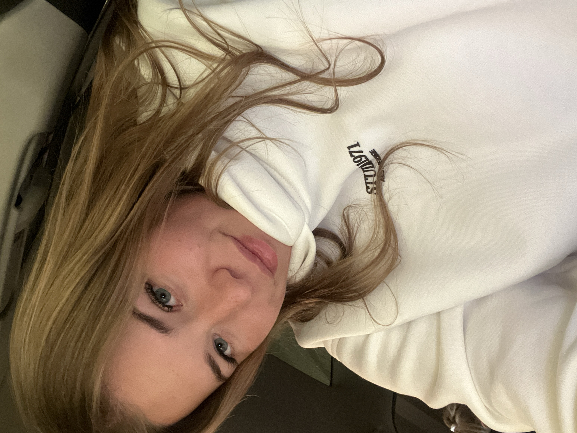 The image displays a close-up, angled portrait of a young adult female. She possesses long, wavy, light brown hair, which is parted on her left and cascades over her shoulders and the front of her torso. Her face shows light-toned skin, with prominent cheekbones, light blue-green eyes, and defined eyebrows. Her lips are closed, forming a slight smile. She is wearing a white high-neck hooded sweatshirt, featuring the black text "STUDIO1971" centrally on the chest. The background is dark, out-of-focus, and indistinct, suggesting an interior environment with low light conditions or dark surfaces, possibly within a vehicle or a room. No specific visual elements within the image confirm the specified location as Budva, Montenegro; this detail is provided externally.