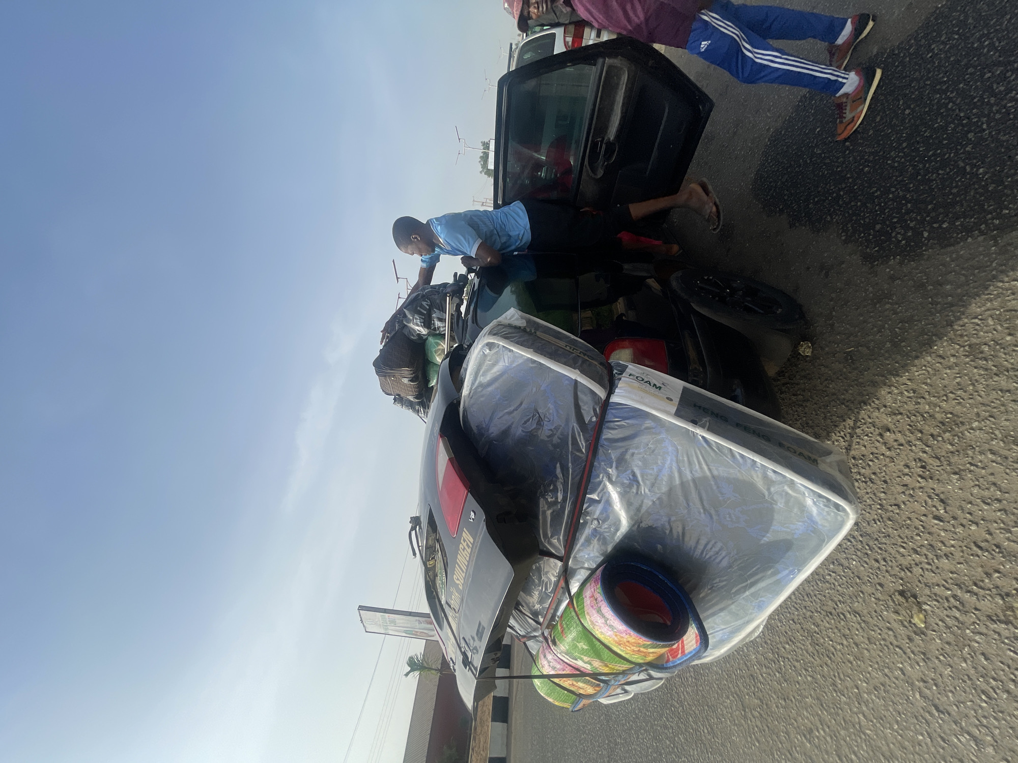 The image shows a black sedan overloaded with items on its roof, secured with straps. A man in a light blue shirt is positioned on the roof, appearing to adjust or secure the load, which includes luggage, a mattress still wrapped in plastic, and rolled-up mats. Another person is visible standing near the open car door. The setting is a paved road with a building or structure visible in the background, identified as being in Jos, Nigeria, based on the license plate details.