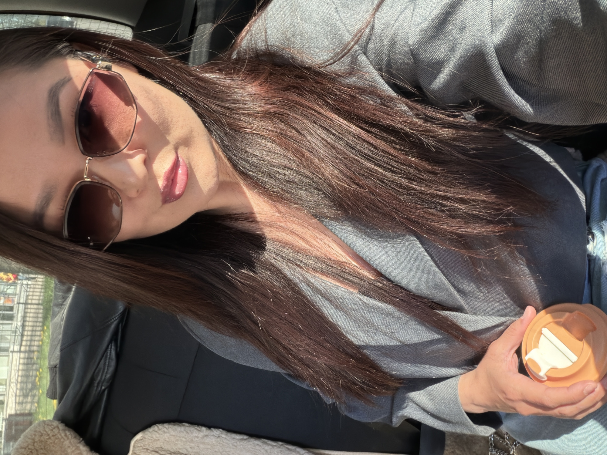 A person is sitting in a car, holding a light orange travel mug with a white lid in their right hand. The person is wearing sunglasses and a grey jacket, with long brown hair. The background shows a blurry view of a city street.
