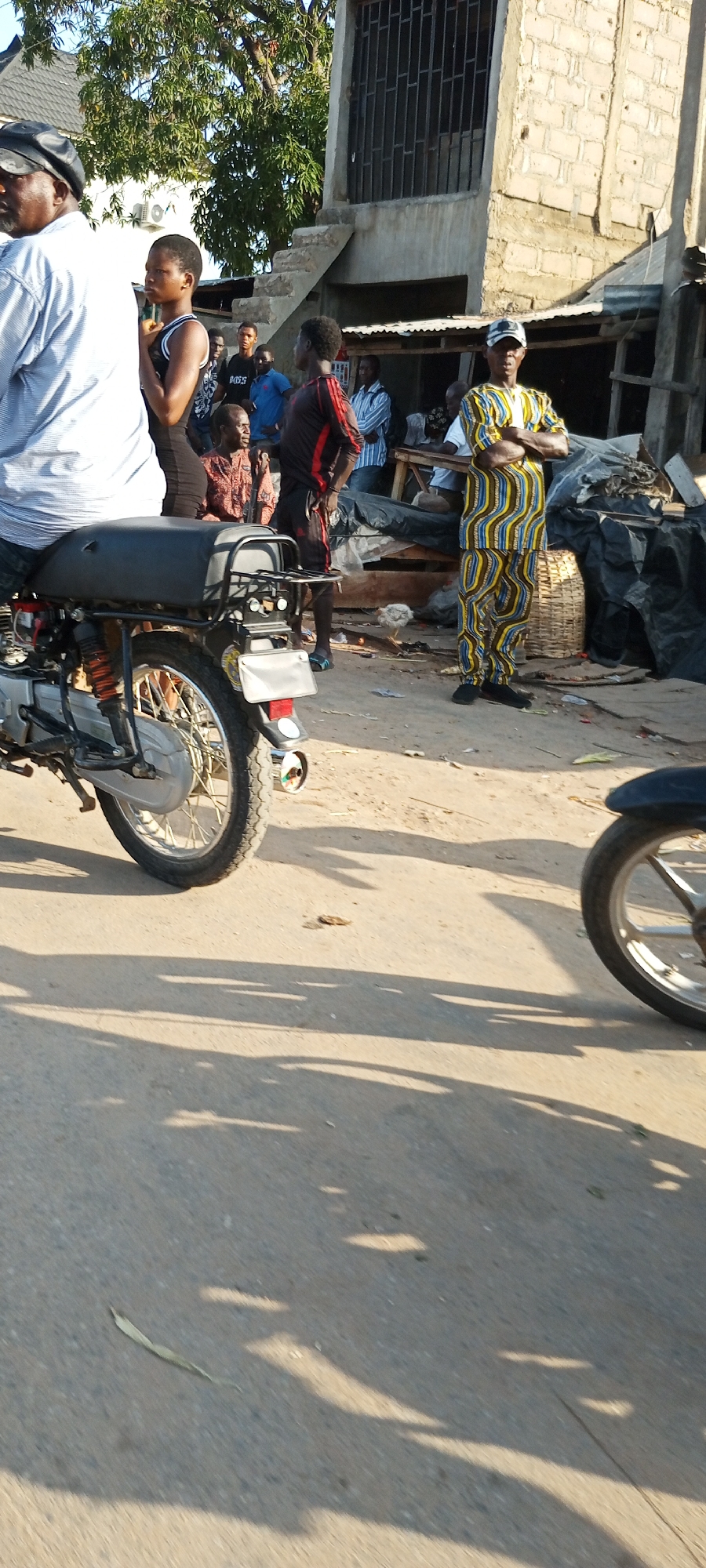 The image captures a street scene in Zango-ndaji, Nigeria. Several people are present, including a man on a motorcycle, a woman in a tank top, a man in a red and black tracksuit, a man in patterned clothing with arms crossed, and a group of people standing and sitting near a building. There are two motorcycles visible in the scene. The environment includes a building with a staircase leading to an upper level, and various objects such as a basket, debris, and what appears to be goods for sale. The street is paved, and shadows from trees or buildings are cast across the surface.