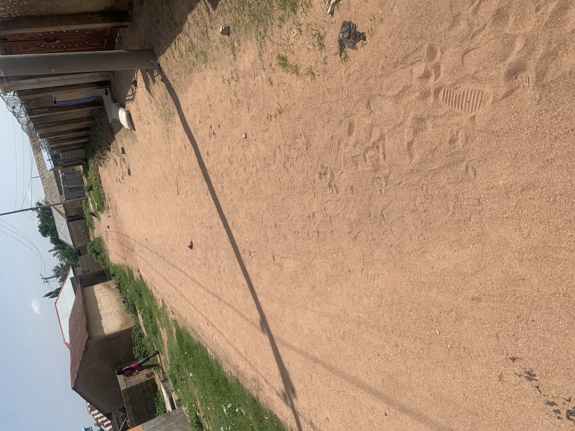 This media file depicts an outdoor scene in Jalingo, Nigeria, on a clear day, likely in the late morning or early afternoon given the long shadows cast by the pole and overhead wires. The ground is predominantly a sandy, reddish-brown dirt path, with some sparse green grass growing along the edges and scattered debris.
In the background, there are buildings with corrugated metal roofs, some appearing to be under construction or partially finished. A tall, slender pole stands near the center of the frame, with overhead electrical wires stretching across the image. Along the left side of the path, a wall topped with barbed wire is visible, suggesting a boundary or security measure.
A single person, a young male wearing a red shirt and dark pants, is standing near a building on the left side of the path, facing away from the camera. The overall impression is a residential or semi-urban area, characterized by its unpaved road and simple structures. The sandy ground shows footprints, indicating recent activity. The lighting suggests bright sunlight with a clear sky. No visible text is present in the image.
