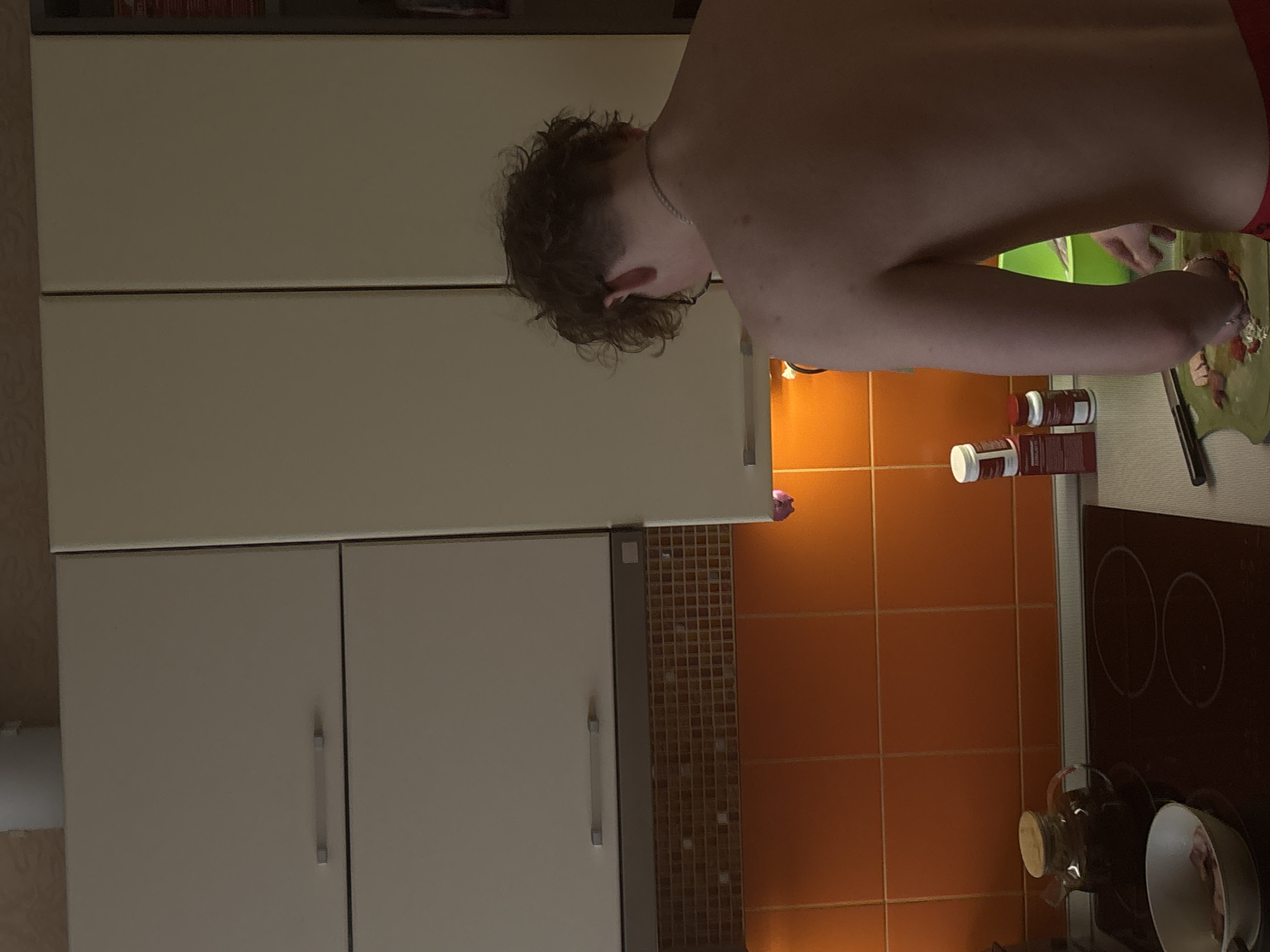The image shows the interior of a kitchen. A shirtless person is standing at a counter preparing food. Their head and upper torso are visible. They have curly brown hair and are wearing a necklace. They are using a knife and cutting board.
The kitchen has cream-colored cabinets, a stove with two burners, and orange tiled walls. There are spice containers on the counter. A glass teapot and white bowl are on the stove.
There is no concrete evidence of a precise location of the kitchen being in Perm, Russia.