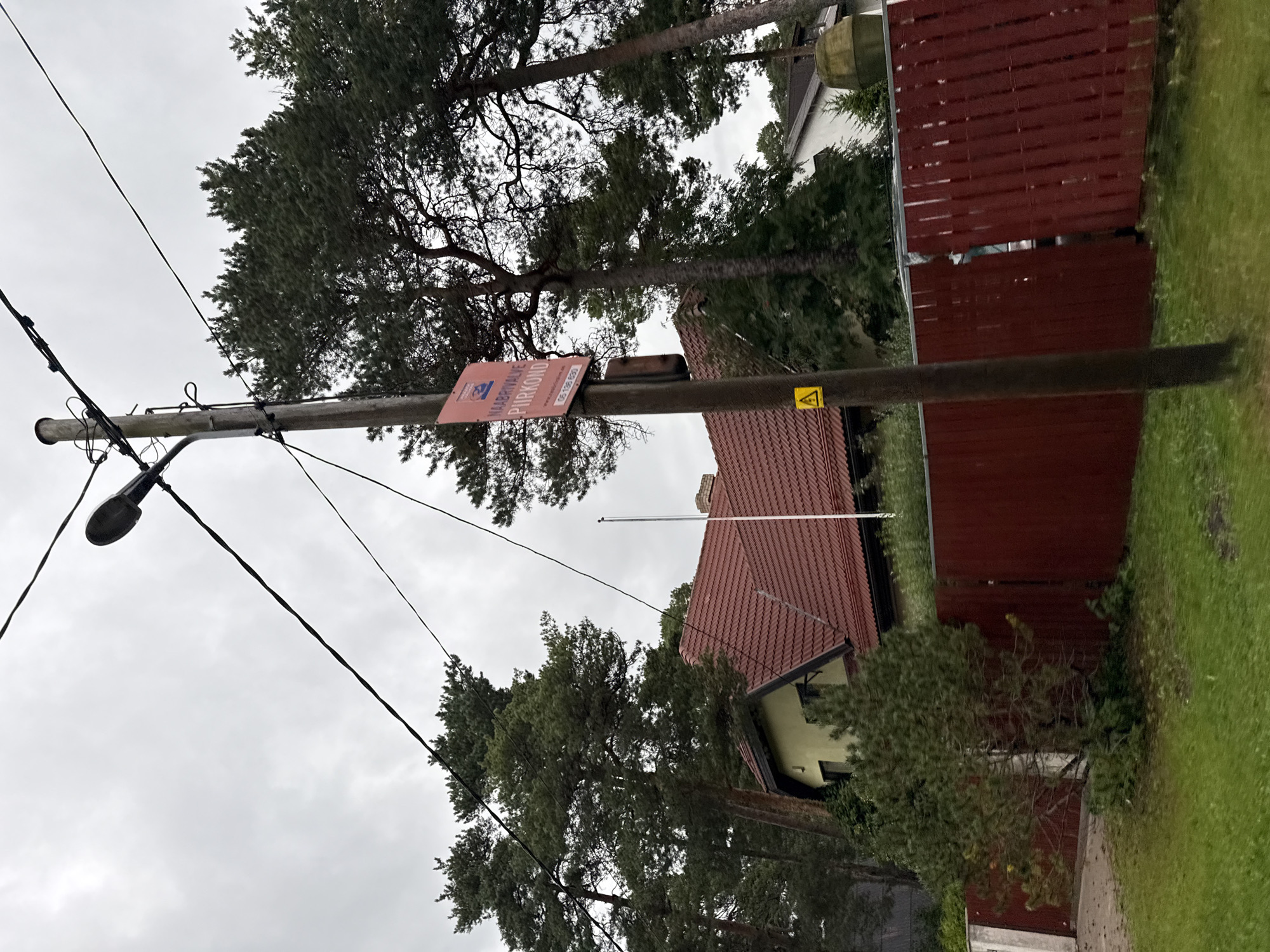 The image shows a residential street in Tallinn, Estonia, with a wooden utility pole in the foreground. The pole is adorned with a red sign that reads "NAABRIVALVE PIIRKOND" and a website and phone number. A yellow triangular warning sign with a lightning bolt is also attached to the pole. Behind the pole, a red wooden fence encloses a property with a house that has a red tile roof and yellow walls. Large pine trees are visible behind the house and fence, and the sky is overcast. The ground is covered in green grass. The overall scene suggests a quiet, suburban neighborhood on a cloudy day.