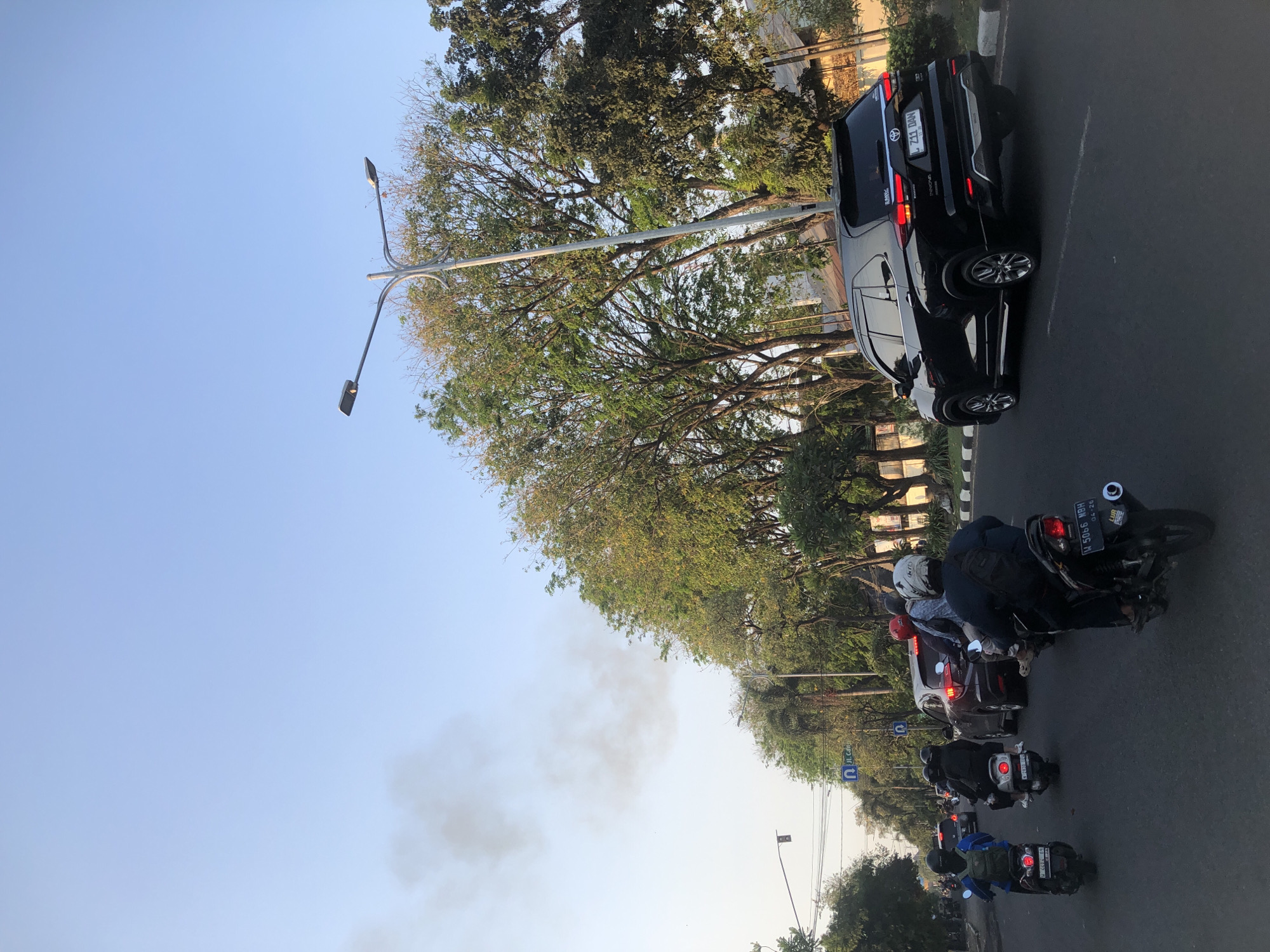 The image depicts a street scene in Candi, Indonesia, with traffic flowing under a clear blue sky. A black Toyota Innova is prominent in the foreground, moving along the road. Several motorcycles are also present, some carrying passengers, indicating typical urban transportation. Tall trees line the street, their leaves displaying a mix of green and autumn colors. A lamppost stands tall, illuminating the road. In the background, a plume of smoke rises into the sky, possibly from a distant fire or industrial activity. The overall atmosphere suggests a busy day in a tropical urban environment, with a hint of an environmental concern due to the smoke.