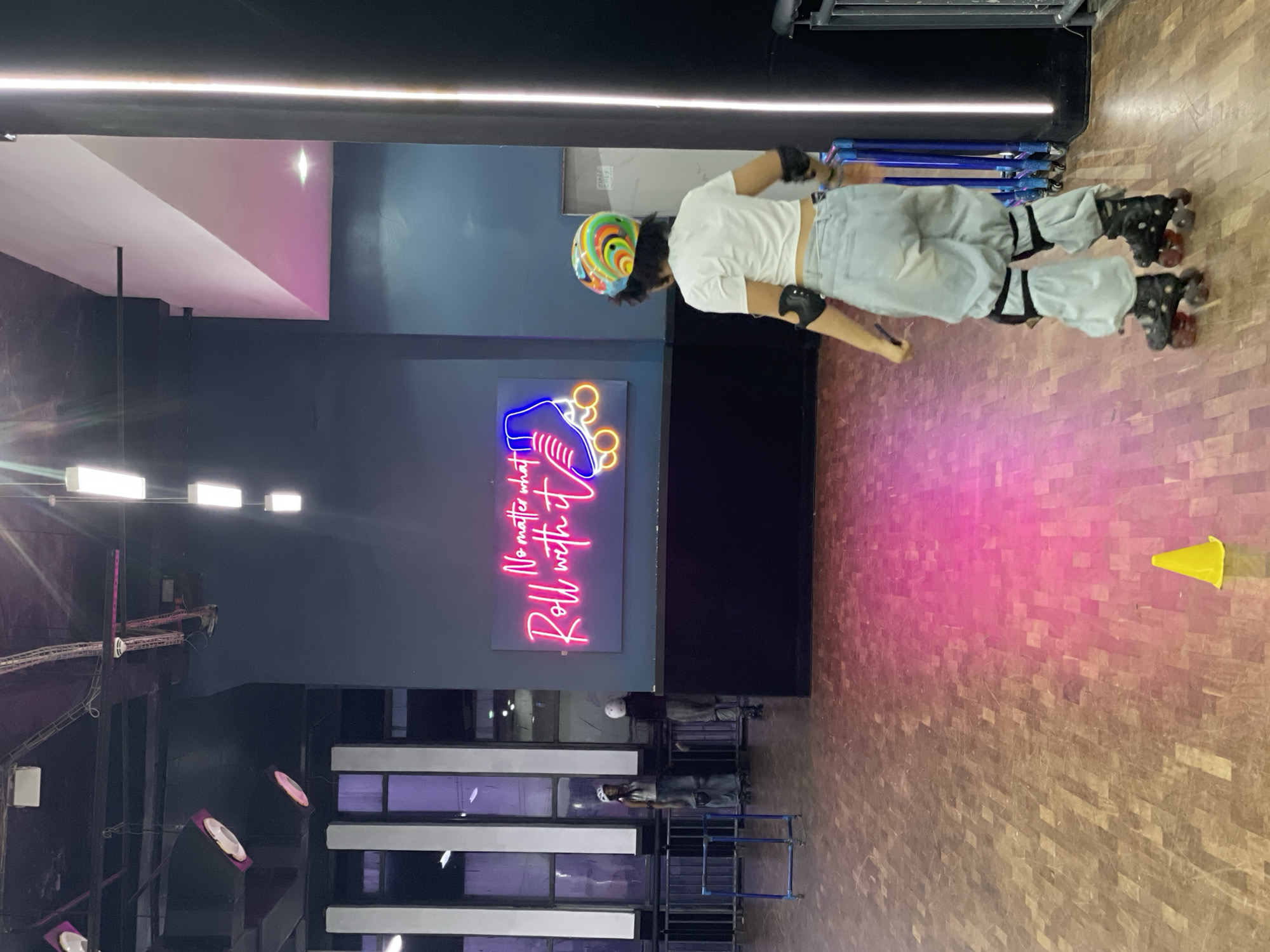 A young person wearing a white t-shirt, light blue baggy pants, a helmet, and roller skates is standing in an indoor roller skating rink. The person is facing away from the camera, looking towards a neon sign on the wall that reads "No matter what Roll with it" and features a roller skate graphic. The floor of the rink is made of wood and is partially illuminated by pink and yellow lighting. In the background, there are other people on roller skates and a metal barrier. The environment appears to be a recreational facility.