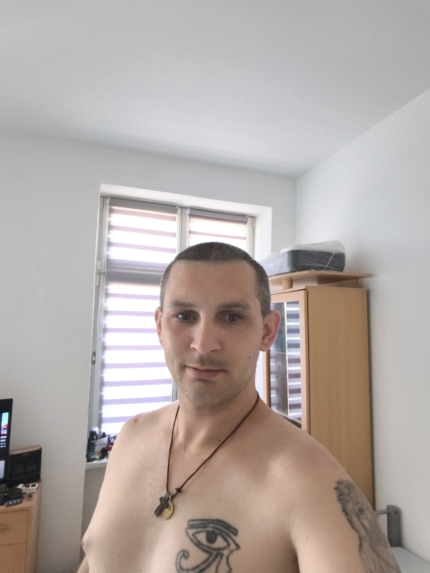 The image depicts a shirtless man in what appears to be a domestic indoor setting. He has a short haircut and is wearing a necklace with a pendant. A tattoo of an eye is visible on his chest, and a partial tattoo is on his right shoulder. He is positioned in front of a window with horizontal blinds and to the right of a wooden cabinet with glass doors. In the background, a television is visible on a wooden dresser to the left. A shelf above the cabinet holds a packaged item. The scene is well-lit, with plain white walls and ceiling. There is no indication of the specific location in the city of Wolgast, Germany.