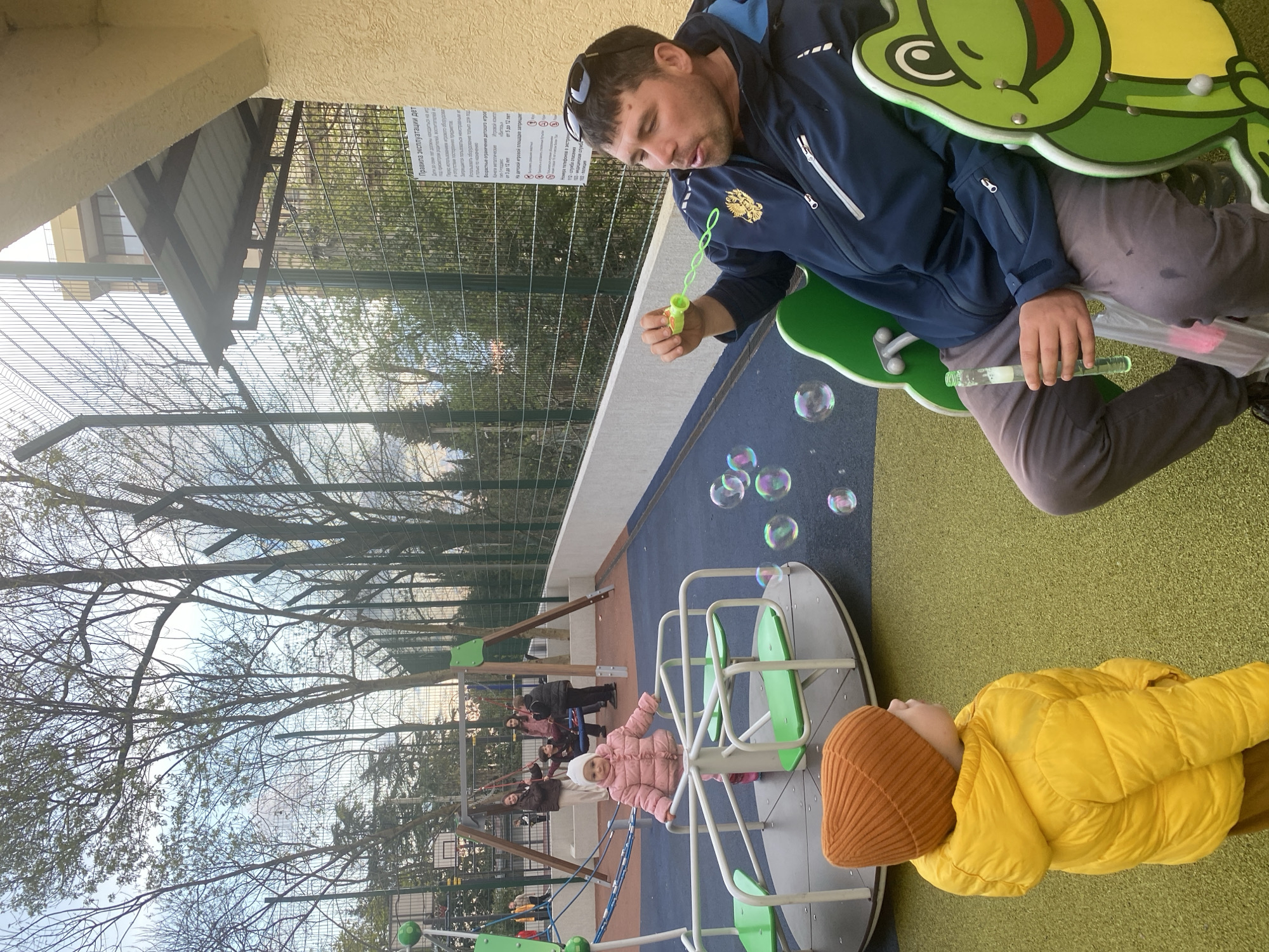 The image depicts a playground scene. A man sits on a green frog-shaped spring rider, blowing bubbles. Nearby, a child in a yellow jacket and brown beanie lies on a green surface. In the background, another child in a pink jacket stands on a gray and green merry-go-round, with additional people visible on swings. Bare trees are visible above a green fence, and a sign written in Russian is affixed to the fence. The man wears a blue jacket and gray pants, and sunglasses rest on his head. The scene is located in the city of Sochi, Russia as evidenced by the Russian state flag on the man's jacket.