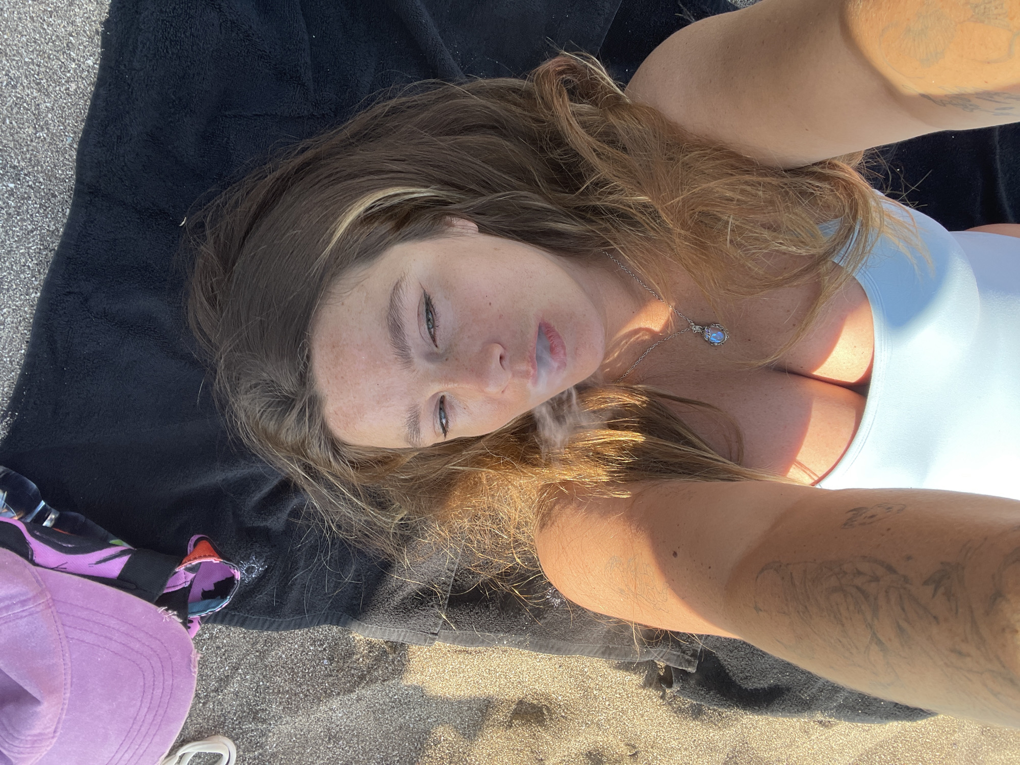 A young woman is lying on her back on a black towel on a sandy beach. She is looking up towards the camera, with her head tilted to her right. A wisp of smoke is visible near her mouth, suggesting she may be smoking. She has long, wavy, auburn hair that falls around her face and shoulders. She is wearing a light blue swimsuit top and a silver necklace with a blue pendant. Her right arm is bent, and her hand is out of frame. She has tattoos on her left arm. To her left, a purple baseball cap and some patterned fabric are visible. The scene appears to be set on a beach, with sand and what looks like a towel on the ground. The lighting suggests it is daytime, possibly late afternoon due to the shadows cast. The woman's expression is relaxed and she appears to be enjoying the moment. There is no visible text in the image.