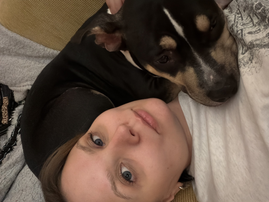 A woman with fair skin and blue eyes lies on her side with a black and brown dog resting its head on her chest. The dog has a white stripe down its forehead. The woman's head is tilted slightly, and she looks directly at the camera. A gray textured surface and a golden-brown surface are visible in the background, suggesting a couch or bed. The overall lighting is soft and natural.