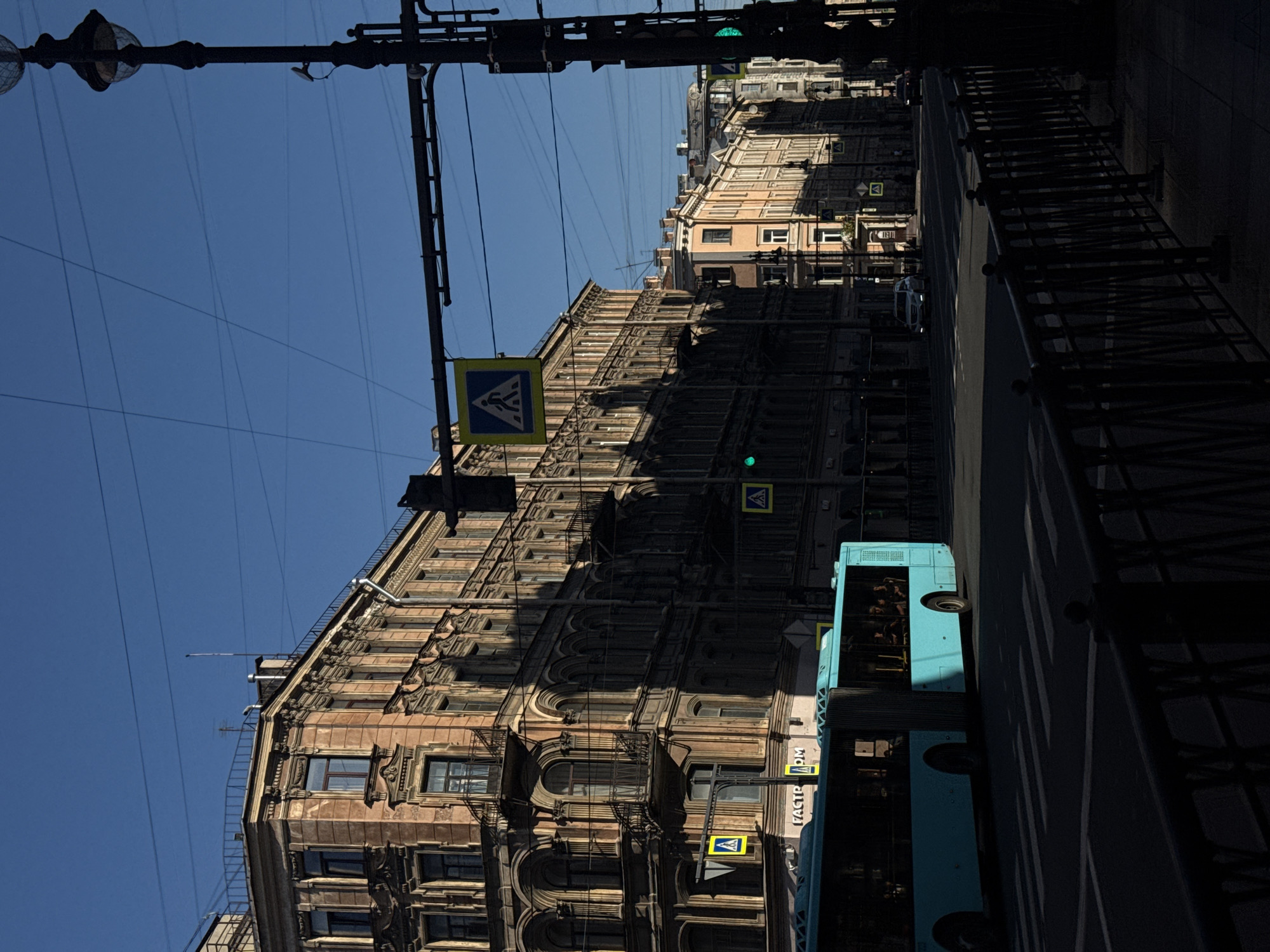 The image depicts a sun-drenched street in Saint Petersburg, Russia, likely during daylight hours with a clear blue sky. The scene is dominated by the architecture of historical buildings lining the street, characterized by ornate facades, arched windows, and balconies. A light blue bus is moving through the frame, crossing a pedestrian crossing. Traffic lights and pedestrian crossing signs are visible, indicating a regulated urban environment. The street appears to be a busy thoroughfare, with power lines crisscrossing the sky and a lamppost on the right side of the image. Shadows cast by the buildings and the bus create a strong contrast, suggesting the sun is at a mid-morning or mid-afternoon angle. The foreground features a dark metal fence, adding to the urban streetscape.