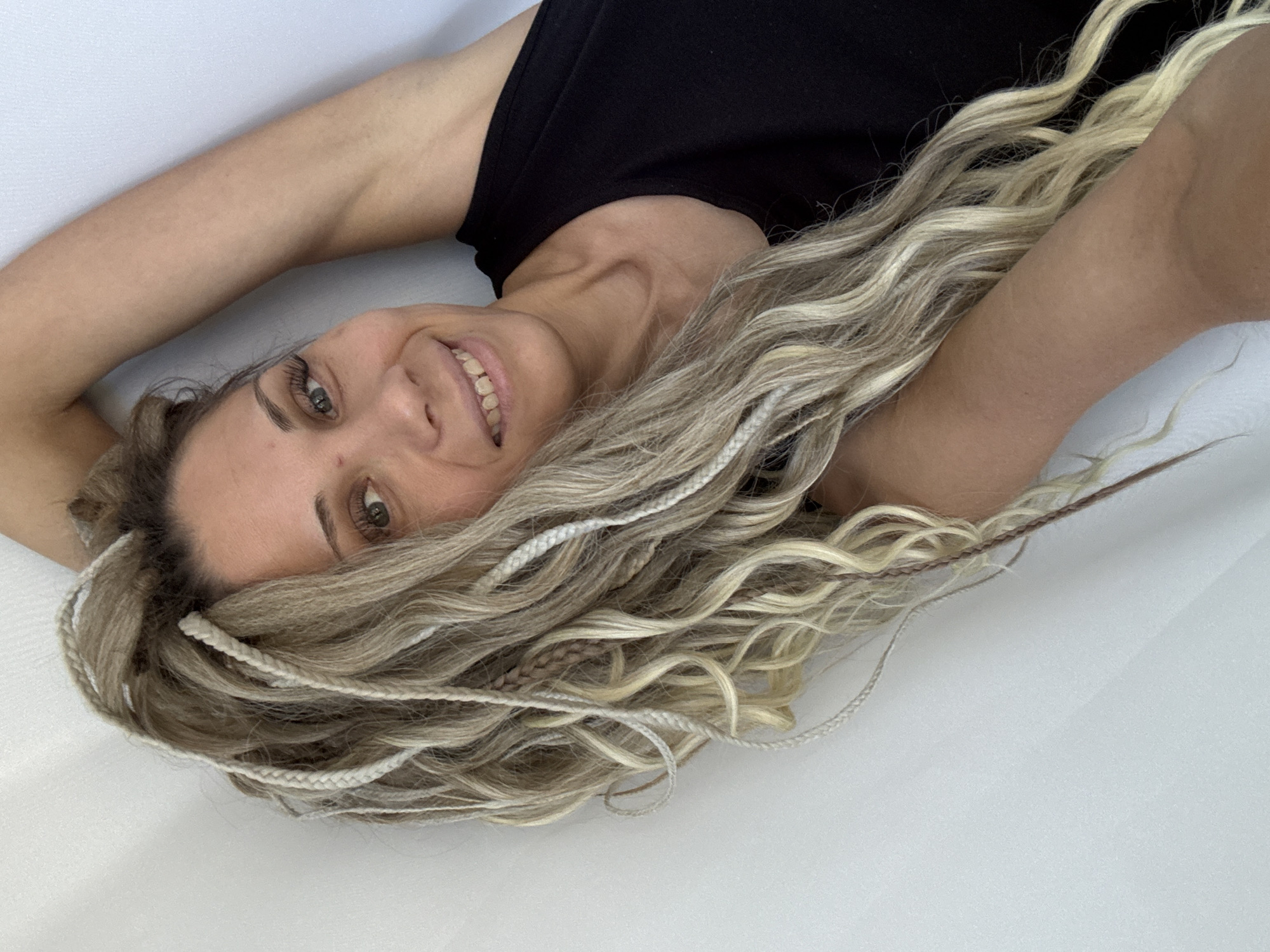 A woman with blonde hair and braids lies on a white surface. She wears a black sleeveless top and smiles at the camera. The location cannot be determined based on the image.