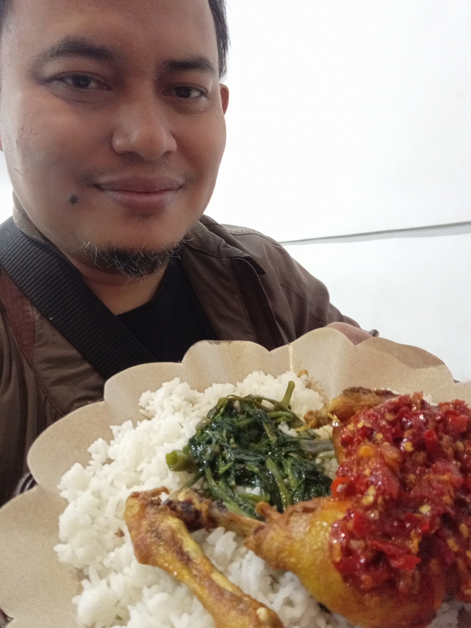 A man, likely in Landungsari, Indonesia, is captured in a close-up, slightly overhead selfie, holding a plate of food. He has a light brown complexion, short dark hair, and a faint goatee and mustache. He is wearing a brown jacket over a dark t-shirt, with a black strap across his chest. The plate, made of disposable brown material, contains white rice, a portion of stir-fried green vegetables, a piece of fried chicken, and a generous serving of red chili sambal. The background is predominantly a plain, light-colored wall.