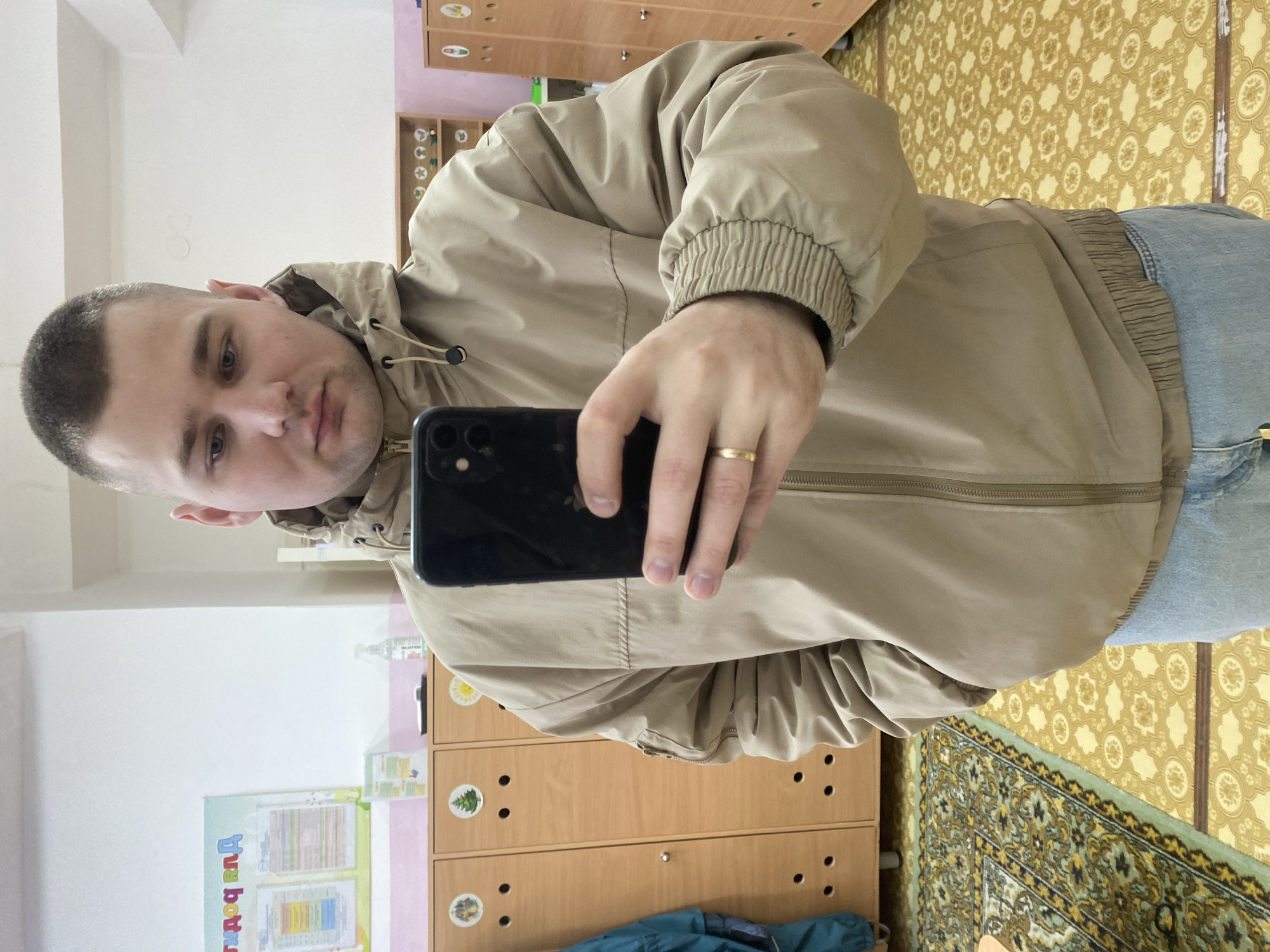 A young adult male, characterized by a short buzz cut and blue eyes, is depicted taking a mirror selfie. He is wearing a beige hooded bomber jacket with a full zipper, ribbed cuffs, and an elasticated waist, paired with light blue jeans. A plain gold wedding band is visible on the ring finger of his left hand. He holds a black smartphone, likely an iPhone, in his right hand, facing its camera towards himself. His left hand is inserted into a jacket pocket. The scene is an interior room of a kindergarten or pre-school located in Chaikovsky, Russia. The background features light wooden locker units, each with individual doors decorated with distinct child-friendly images such as a house, a Christmas tree, and a sun. The walls consist of a white upper section and a light pink lower section. A light blue-green poster with Cyrillic text and charted information is affixed to the wall above the lockers. The floor exhibits two distinct patterns: a traditional patterned rug with green, gold, and brown hues on the left, and a yellow-gold linoleum or vinyl flooring with a repeating geometric pattern on the right. A white spray bottle and other small items are visible on top of the locker units.