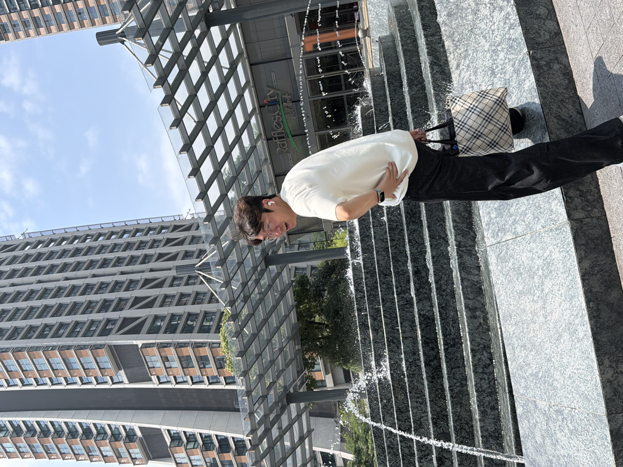 A young East Asian man stands in front of a fountain with tiered stone steps in Yuzhong District, China. He is wearing a white t-shirt, black pants, and white earbuds, and he is carrying a plaid tote bag. He looks over his left shoulder at the camera with a slight smile. Behind him, a building with a glass facade is visible, and the text "Raffles City" is displayed on the building. Tall modern buildings are visible in the background. The sky is bright and clear.