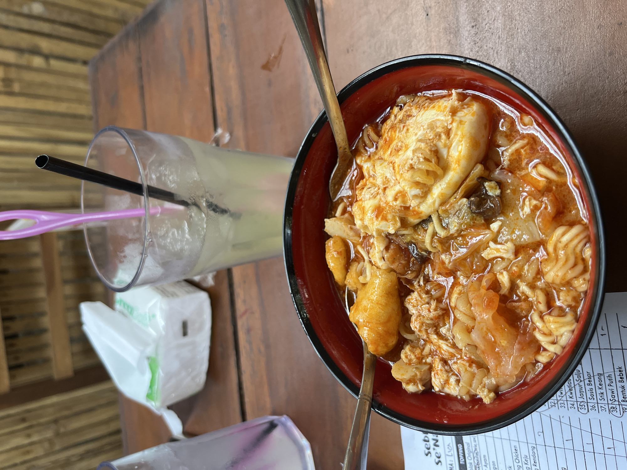 A close-up overhead shot shows a red and black bowl of Indonesian food, possibly noodles or a stew, with various toppings including what appears to be fried egg and meat. Two spoons are placed in the bowl. In the background, a glass of pale yellow liquid with ice and two straws sits on a wooden table. A pack of tissues is also visible. The setting appears to be an outdoor eating area with a bamboo-like wall in the background. The menu on the table indicates the location is in Matesih, Indonesia, and lists food items like "Jamur Salju" and "Bebek".