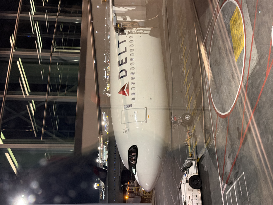 A Delta aircraft is parked at an airport gate. The plane, white with red and blue Delta branding, is connected to a tug vehicle. The terminal building, featuring large glass windows reflecting interior lights, is adjacent to the aircraft. Yellow painted markings on the concrete indicate designated areas for ground support vehicles.
