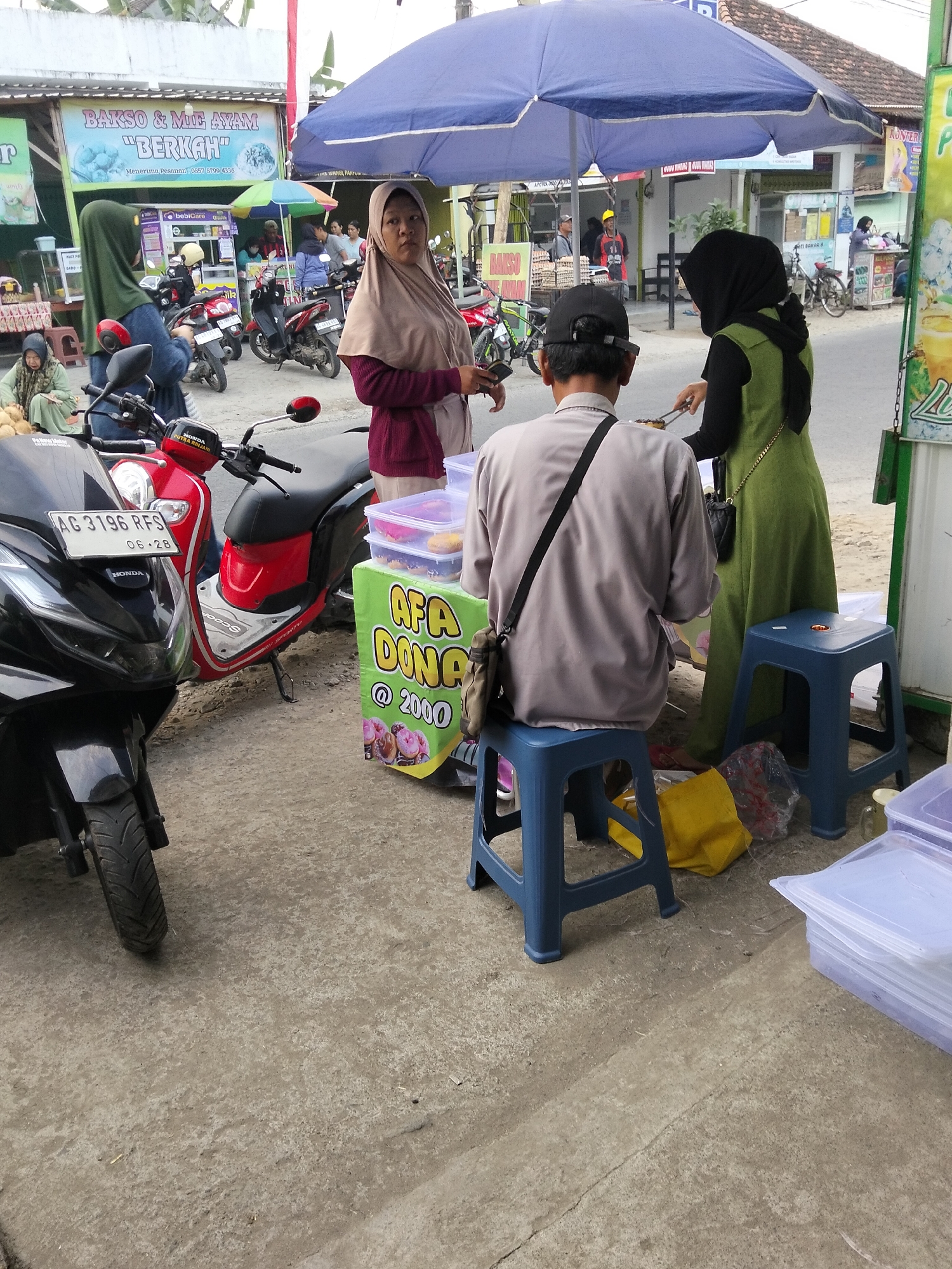 The image depicts a street scene in Gempolan, Indonesia, with a focus on a food stall and its patrons. In the foreground, two scooters are parked, one black and one red. A food stall with a green banner advertising "AFA DONUT @ 2000" is visible, displaying donuts in clear plastic containers. A man wearing a black cap and a grey shirt sits on a blue stool in front of the stall, holding a phone. Another person, a woman in a green hijab and dress, stands behind the stall, seemingly serving food. A woman in a beige hijab and maroon top also stands near the stall, looking towards the viewer. In the background, there are other people, buildings, and parked motorcycles. The overall environment suggests a bustling local marketplace or roadside eatery.